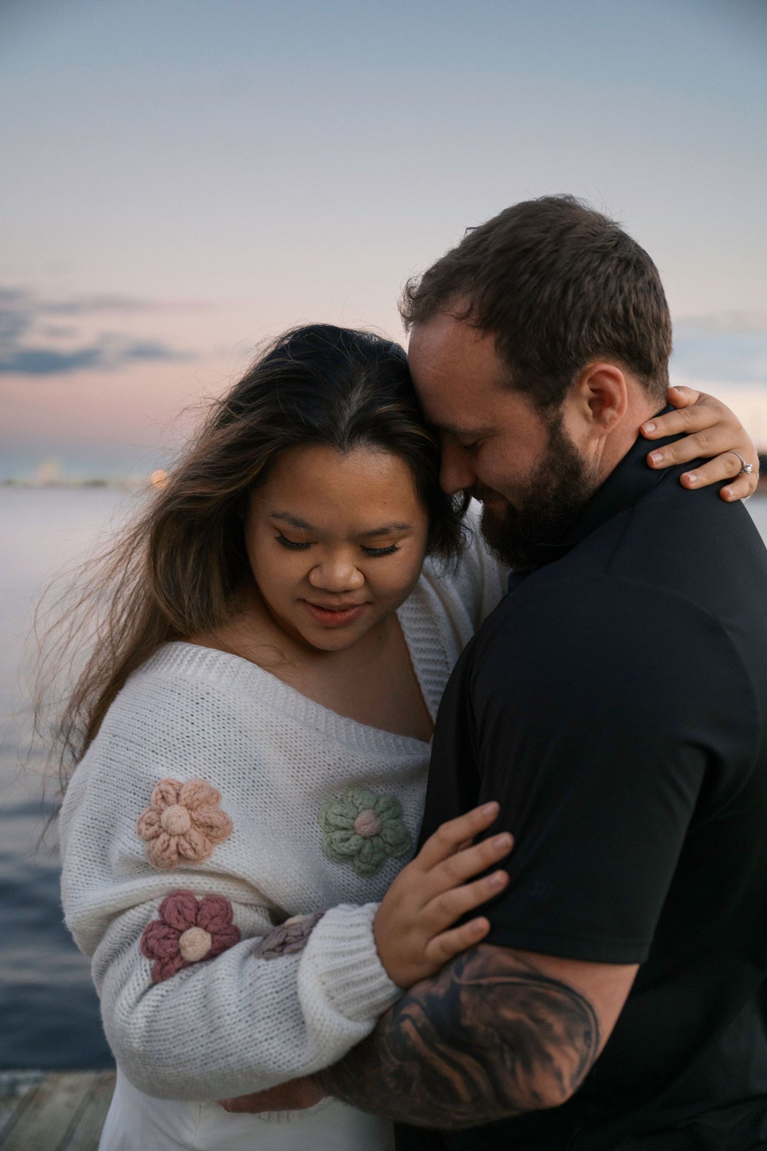 A romantic couple embraces on a dock at sunset with the ocean and pastel sky in the background.