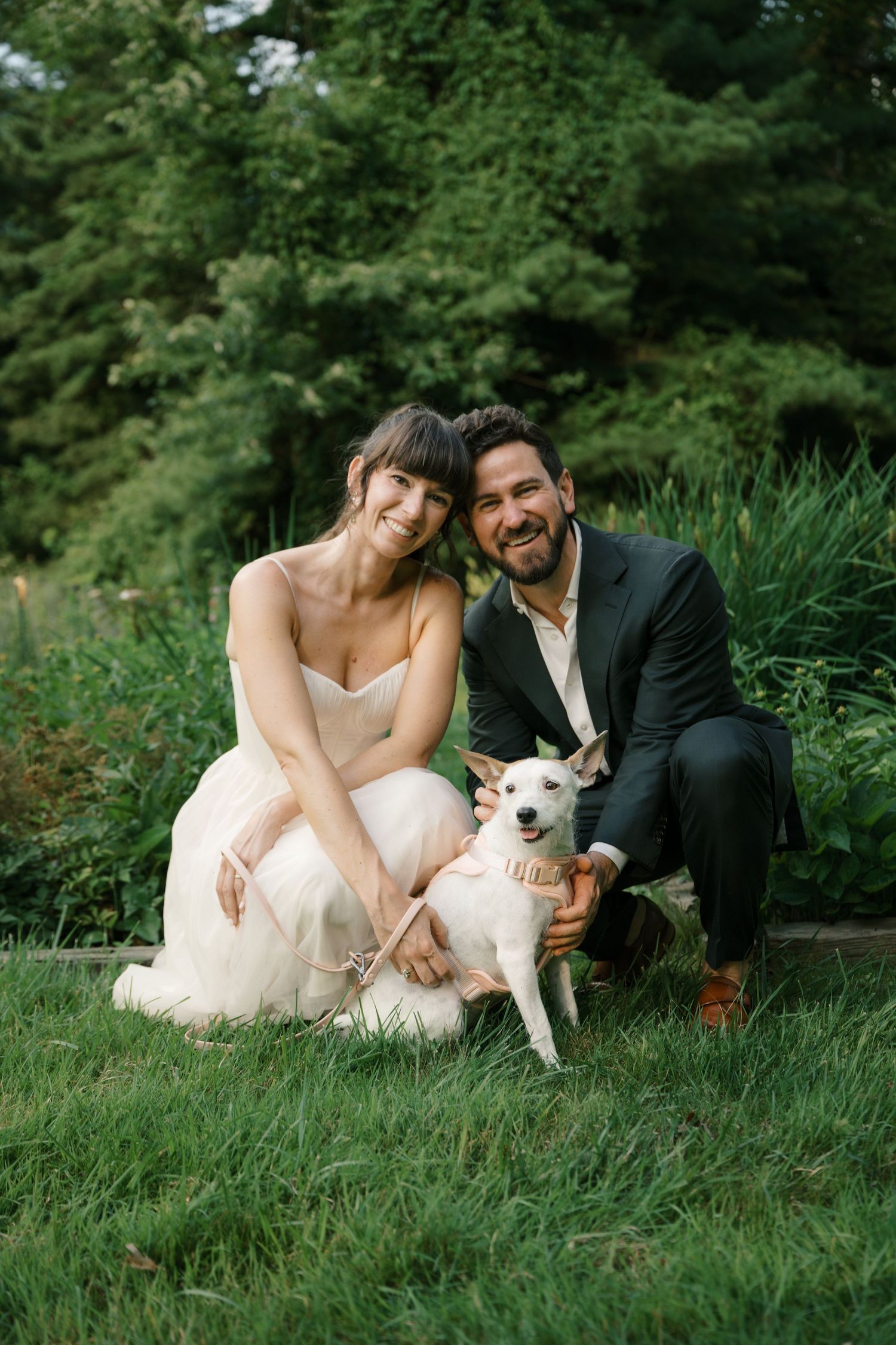Elegantly dressed couple poses with their white dog on grassy hill surrounded by lush forest.