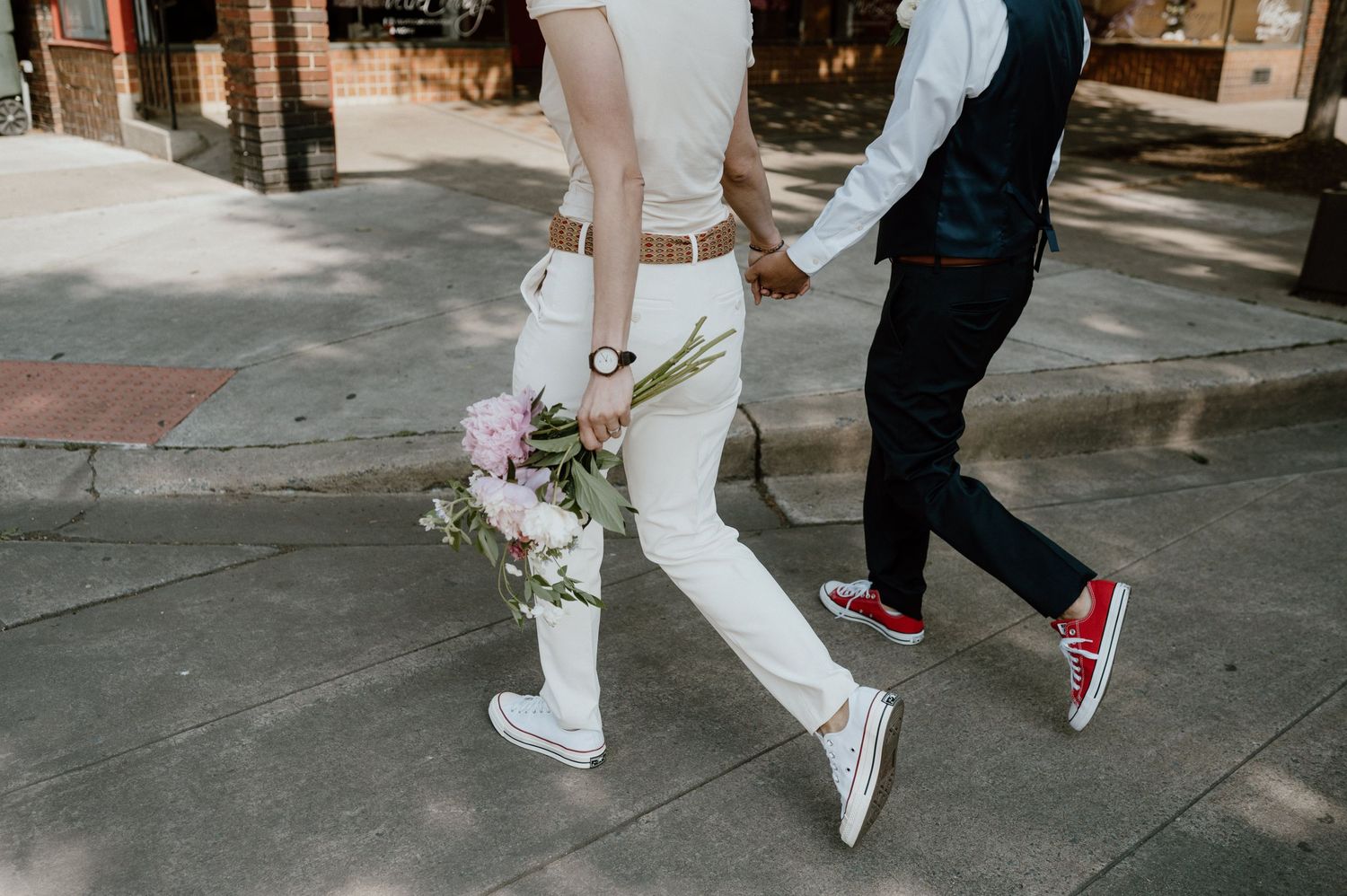 Two people walking hand in hand on a sidewalk, wearing sneakers and formal attire.