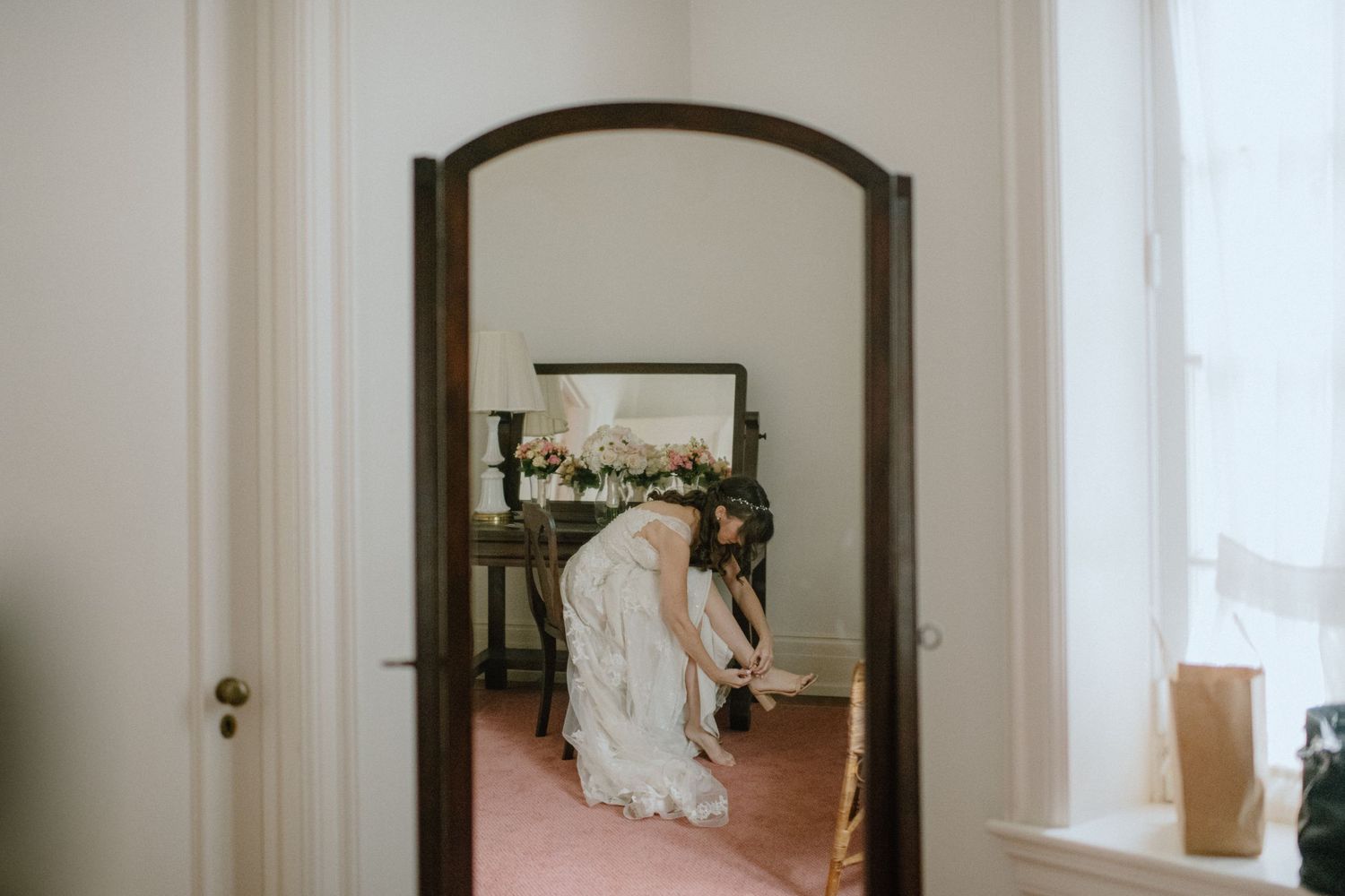 Mirror reflection through an arched doorway showing a wedding dress hanging in a room.