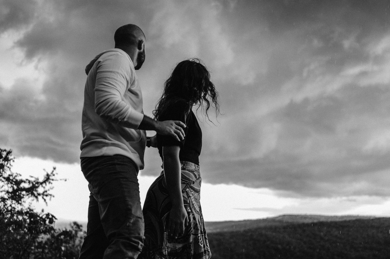 Couple embraces on a hilltop against dramatic stormy skies in black and white.