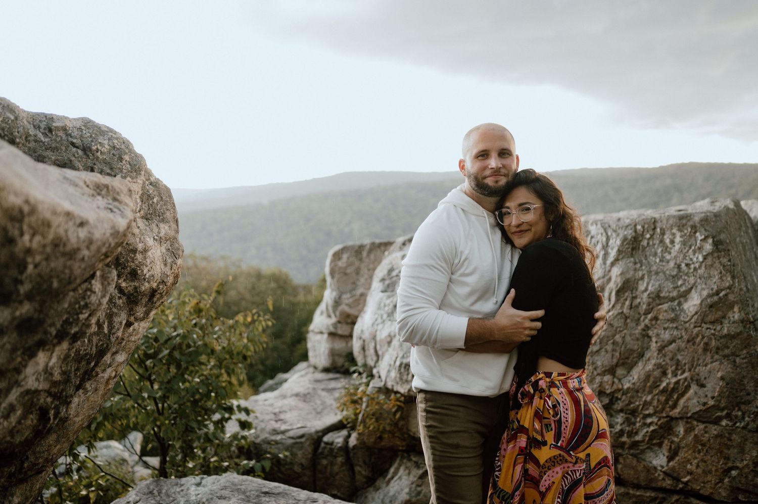 A couple in casual attire embracing while standing among large rocks with mountains in background.
