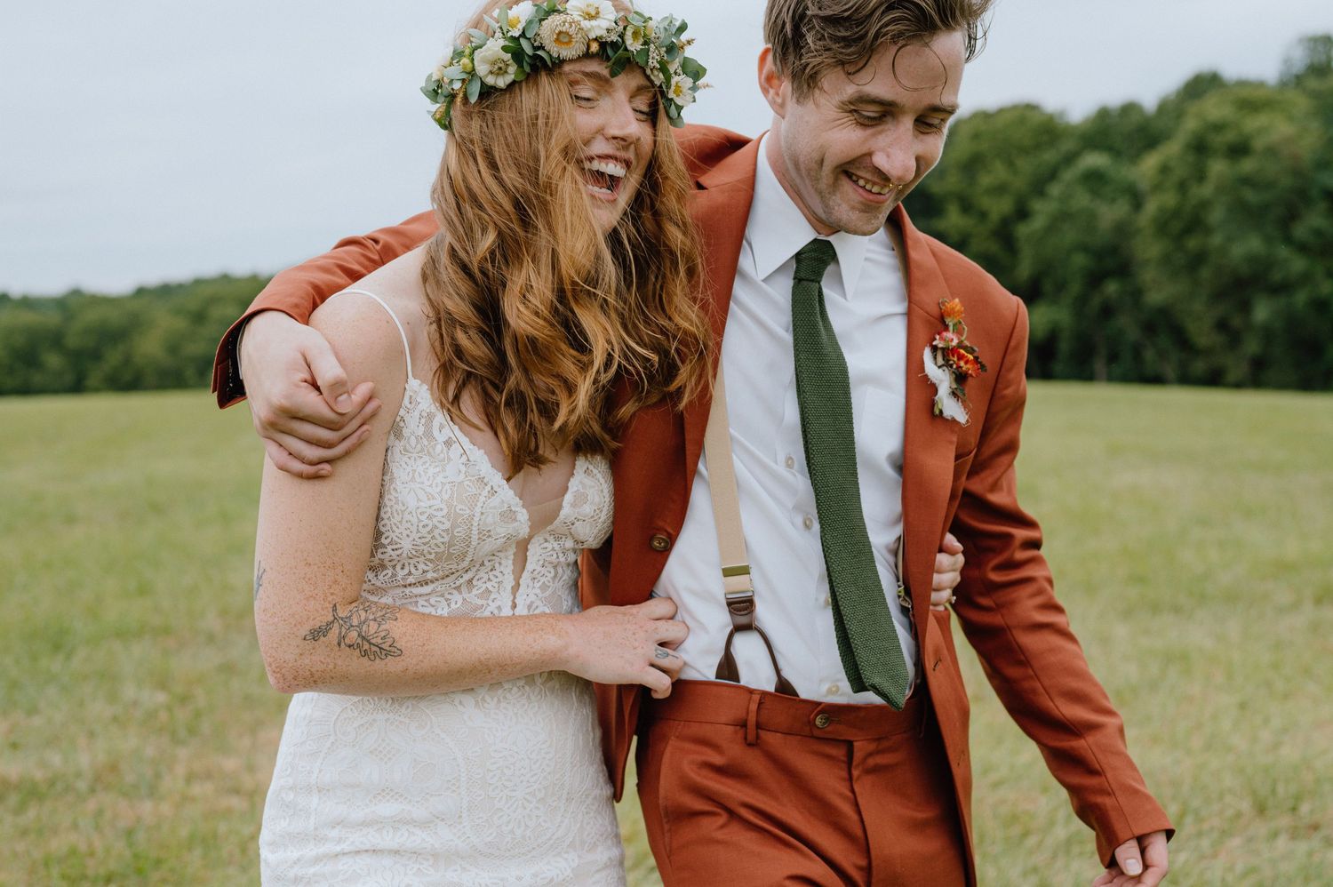 A romantic outdoor wedding photo showing a couple in a lace gown and rusty orange suit laughing together in a field.