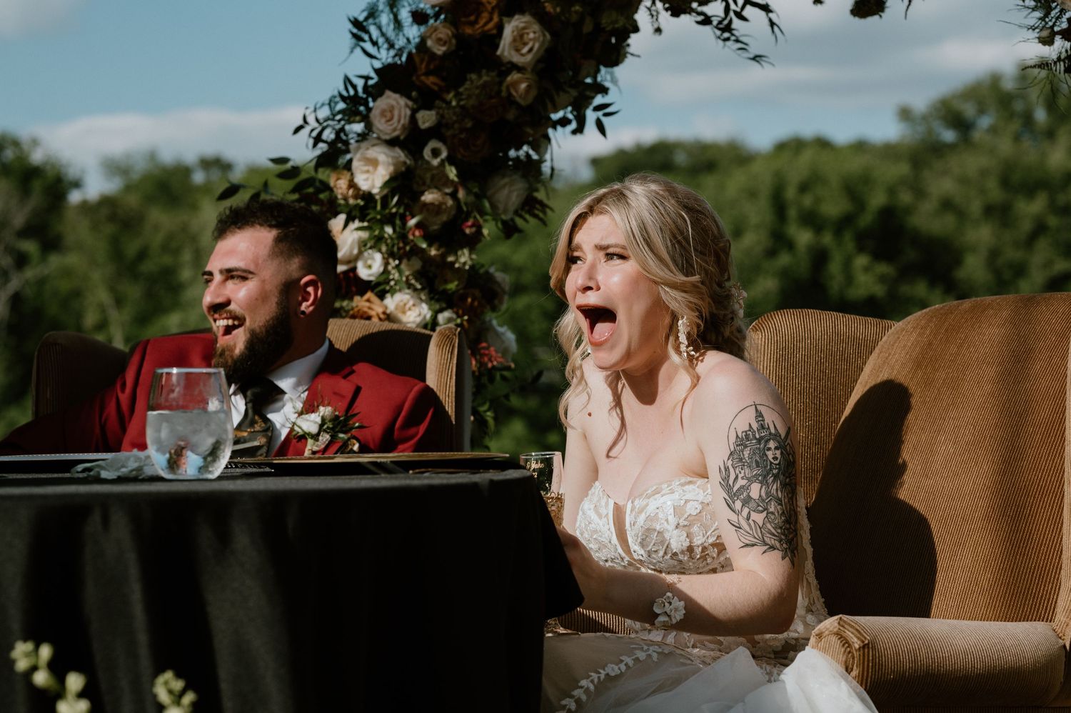 Wedding couple sharing a laugh at their sweetheart table under floral arch.