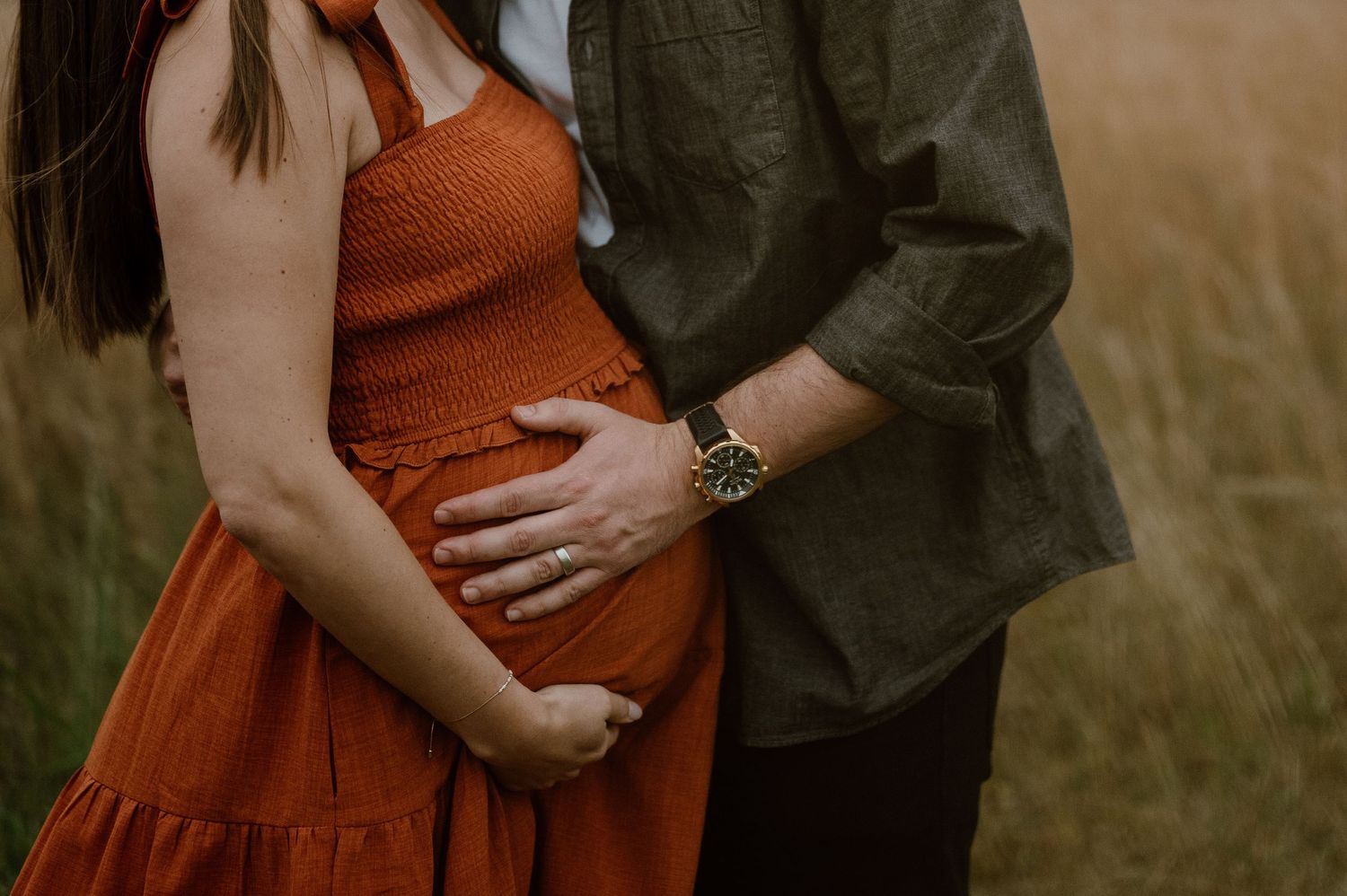 A couple in a field gently holds hands over a pregnant belly wearing an orange dress and dark shirt.