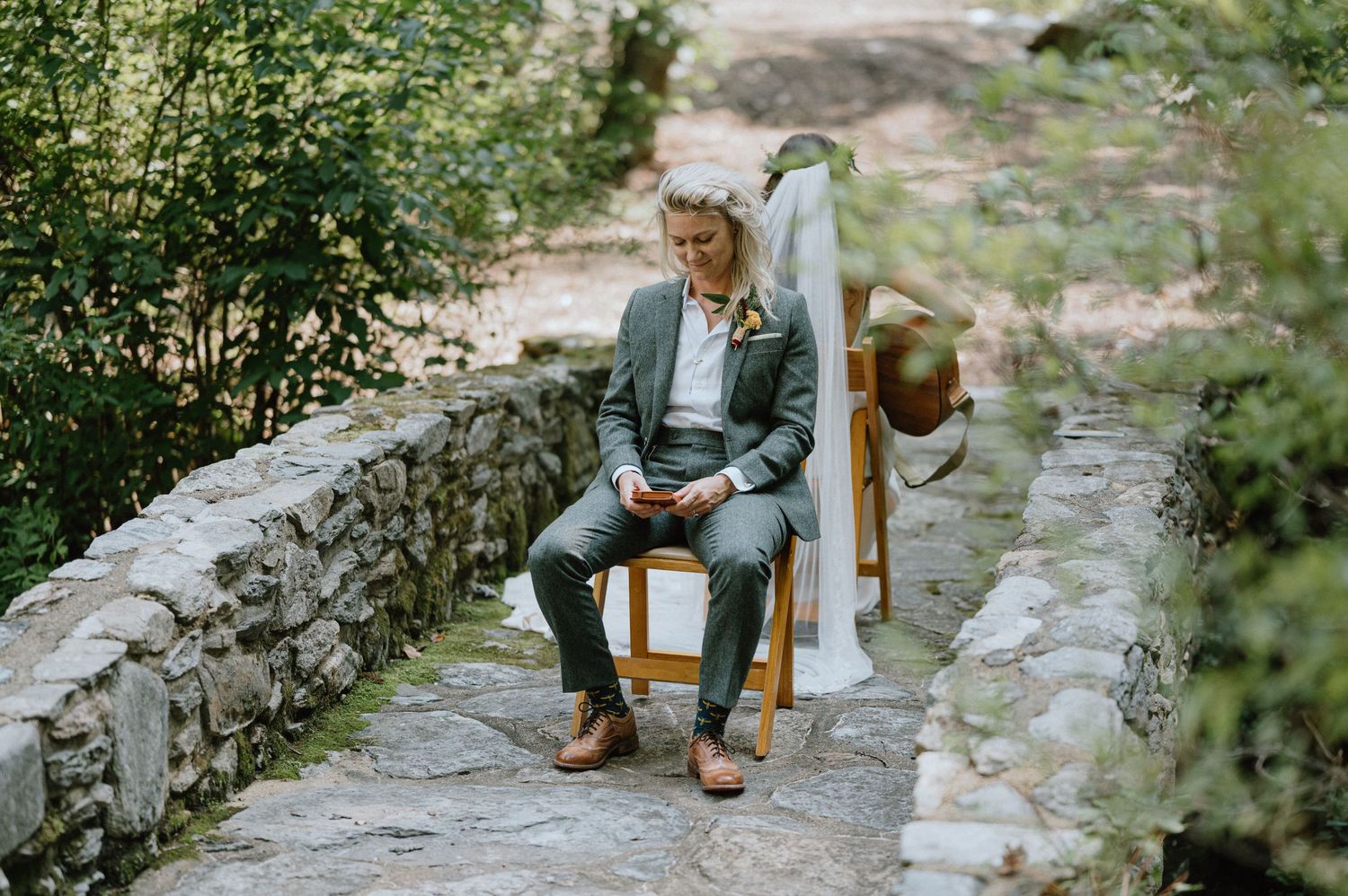 A contemplative figure sits on a wooden chair along a stone-lined garden path surrounded by greenery.