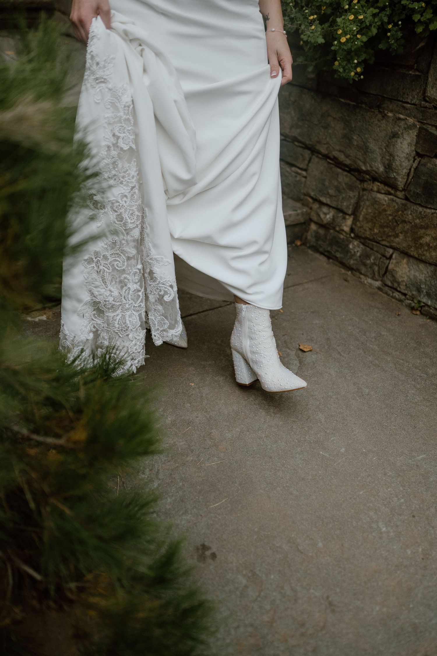 Close-up detail of white wedding dress hem and lace with matching white boots against stone wall and greenery.