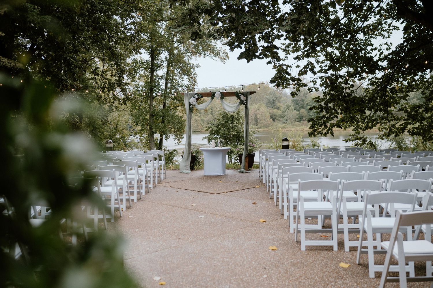 Outdoor wedding ceremony setup with white chairs and wooden arch amongst trees.