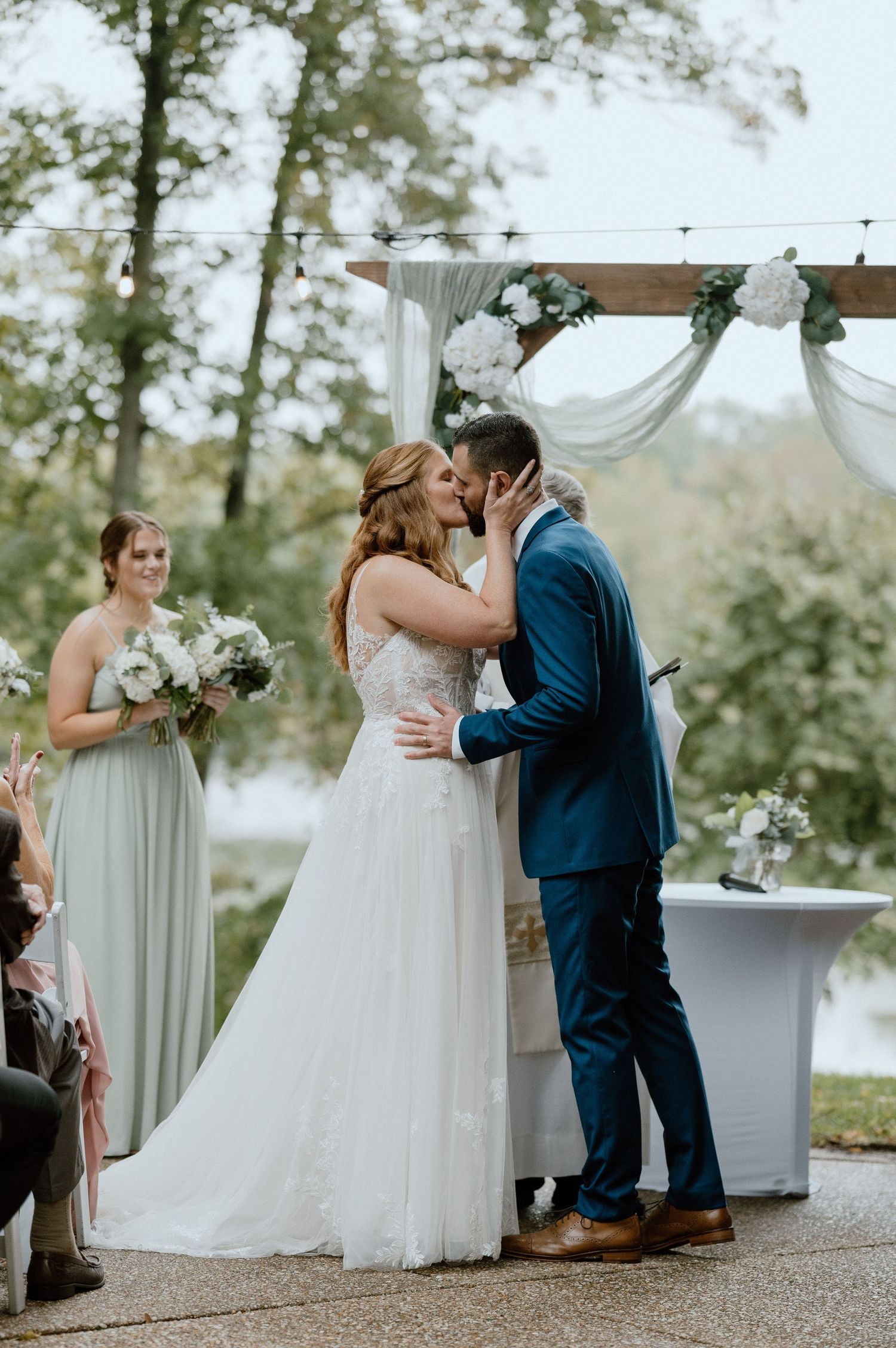 Newlyweds share their first kiss under a white draped arch during an outdoor wedding ceremony by a lake.