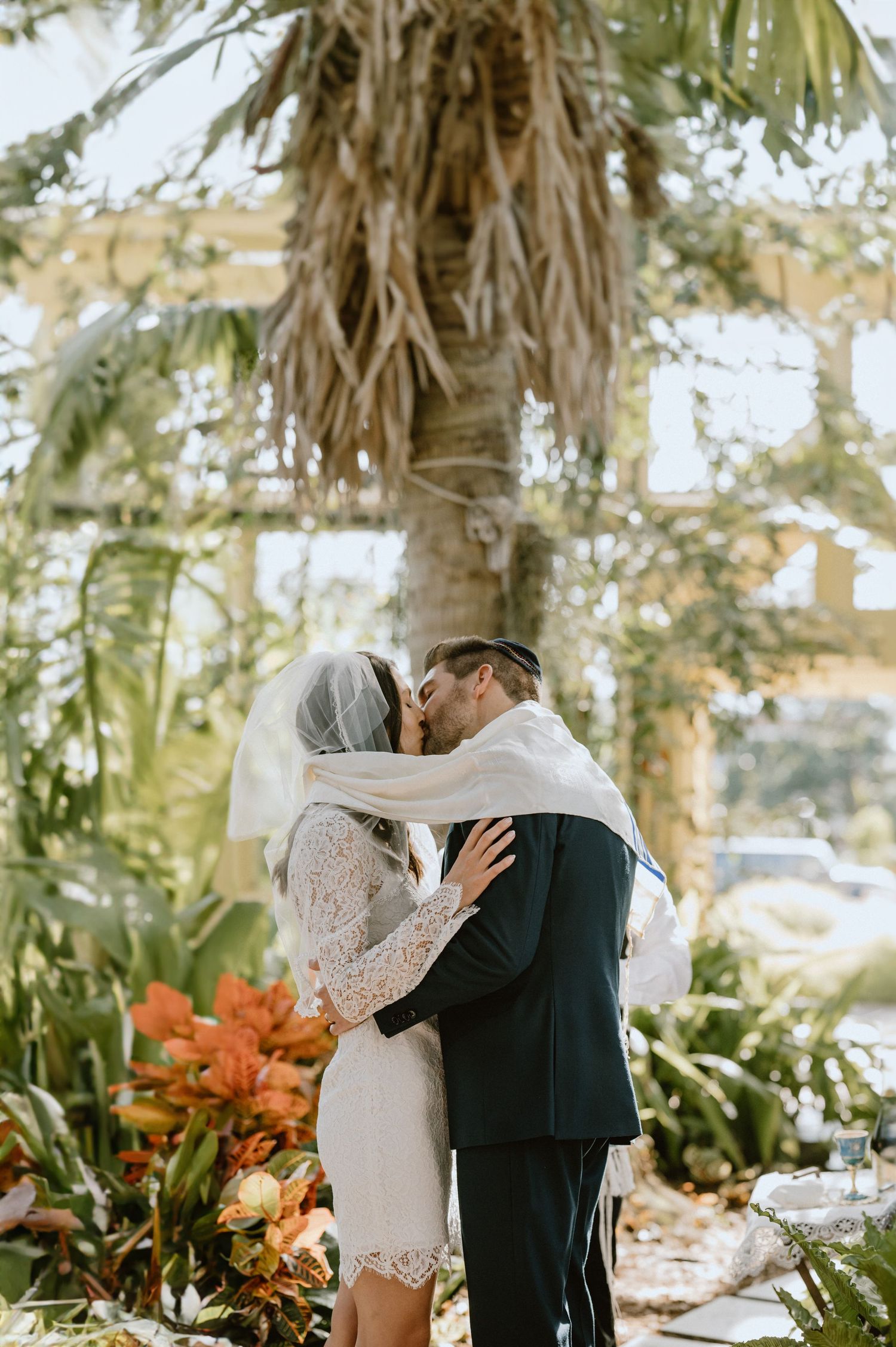 A couple shares their first kiss as newlyweds in a sunlit greenhouse filled with tropical plants.