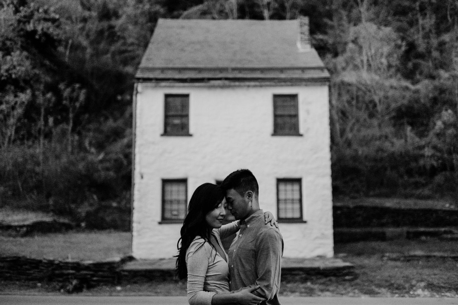 A couple embraces in front of a white historic house in black and white photography.