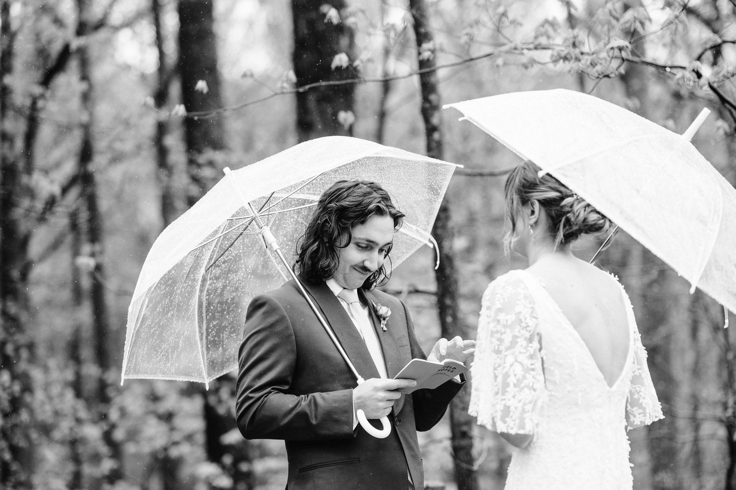 A black and white photo captures a wedding ceremony moment under clear umbrellas in the woods.