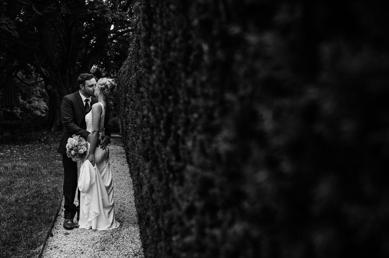 A black and white wedding portrait of a couple framed by garden hedges.