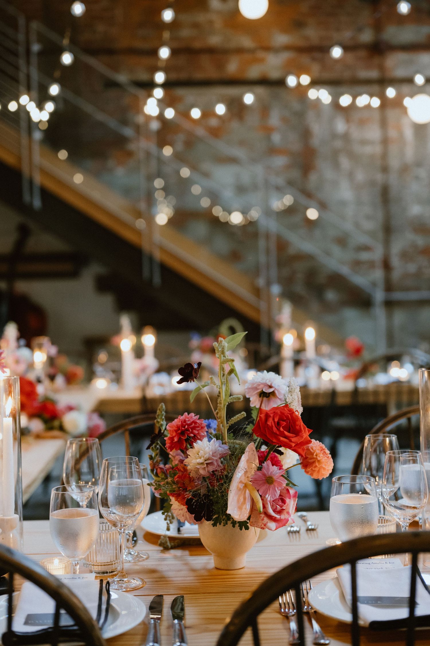 An elegant table setting with colorful floral centerpiece, wine glasses and string lights overhead.