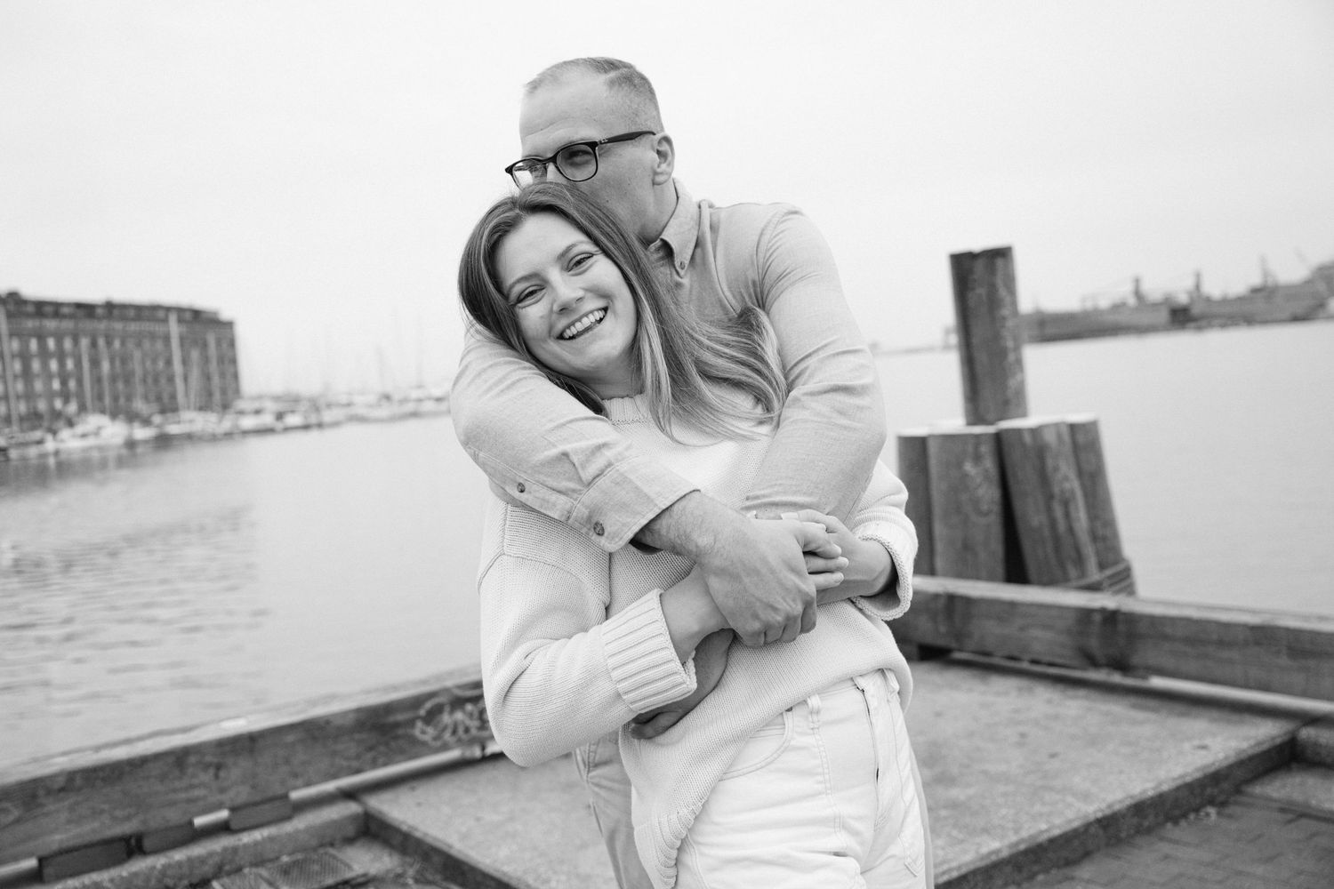 Black and white photo of a couple embracing and smiling on a waterfront dock.