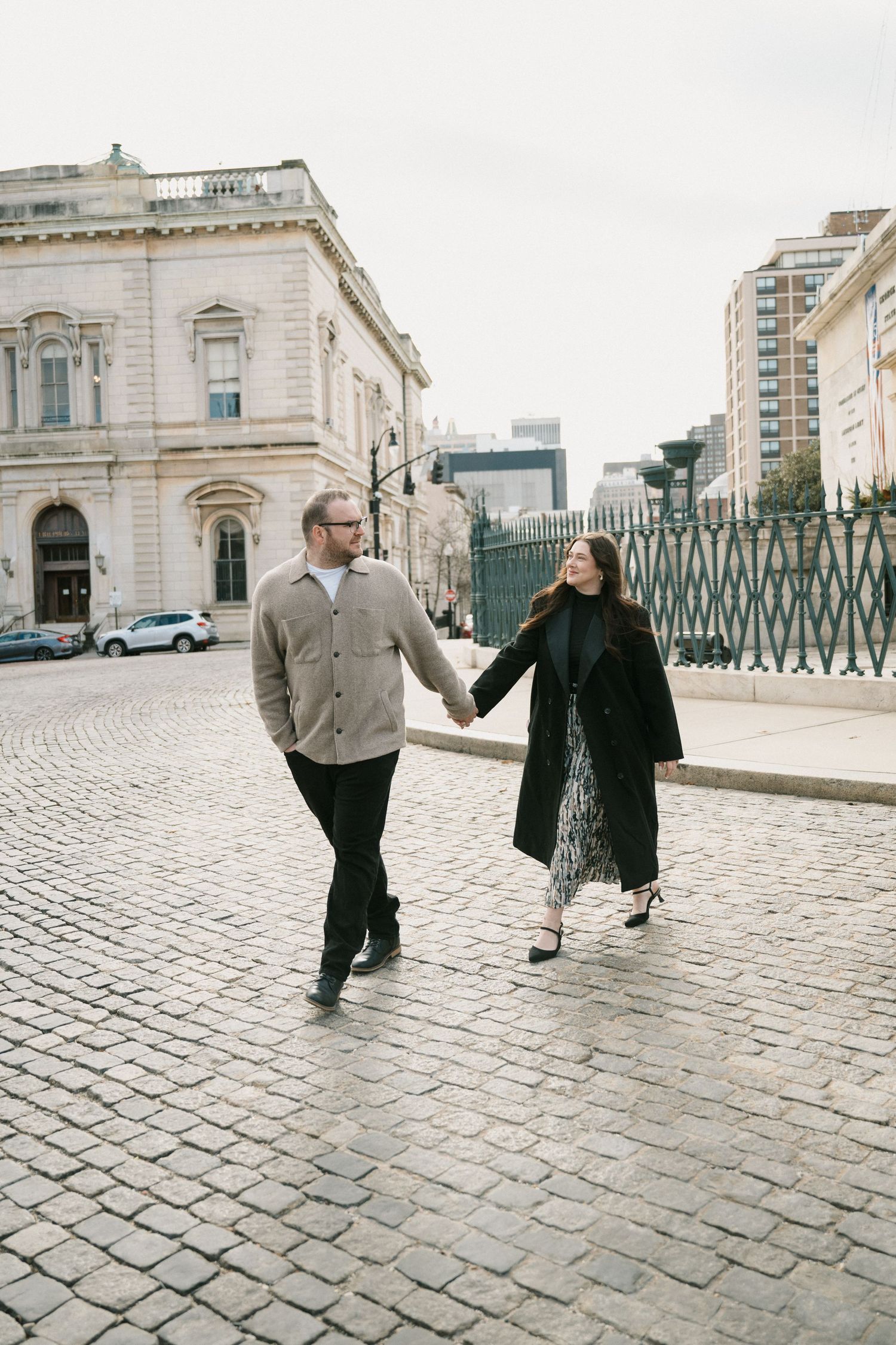Couple holding hands walking on cobblestone street in urban setting, woman in black coat and man in gray jacket.
