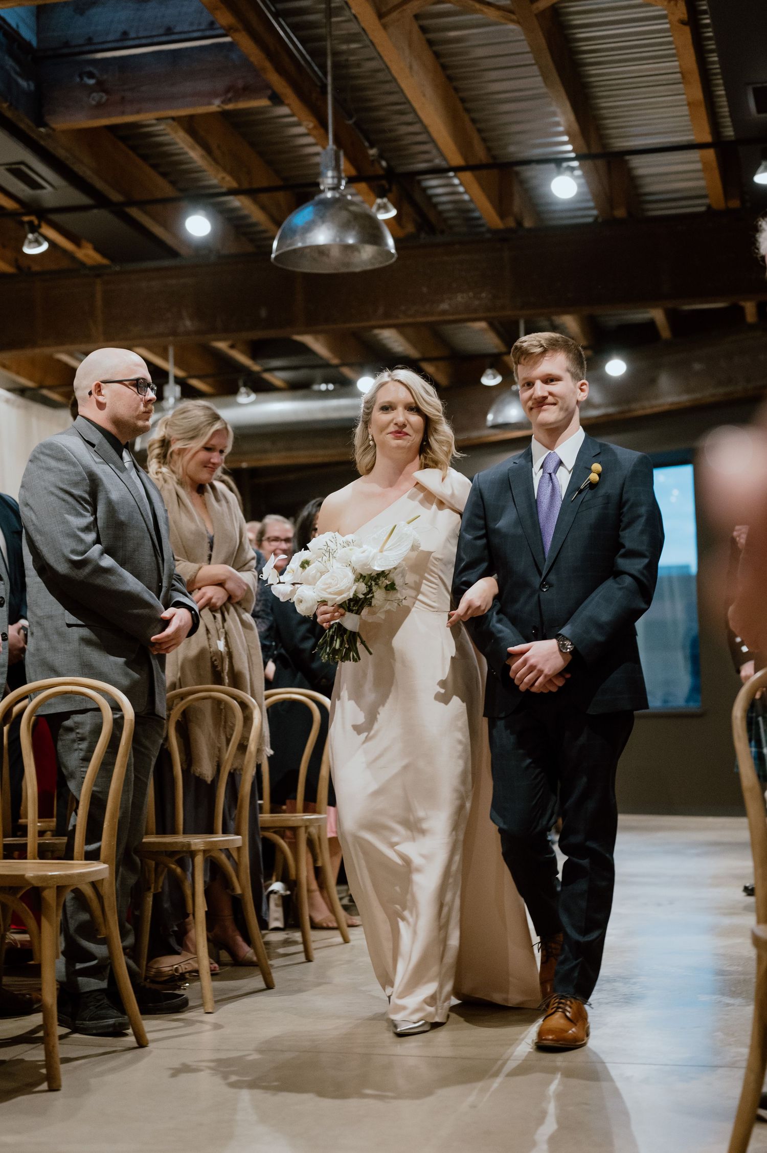 Wedding ceremony in an industrial venue with wooden chairs and pendant lighting.