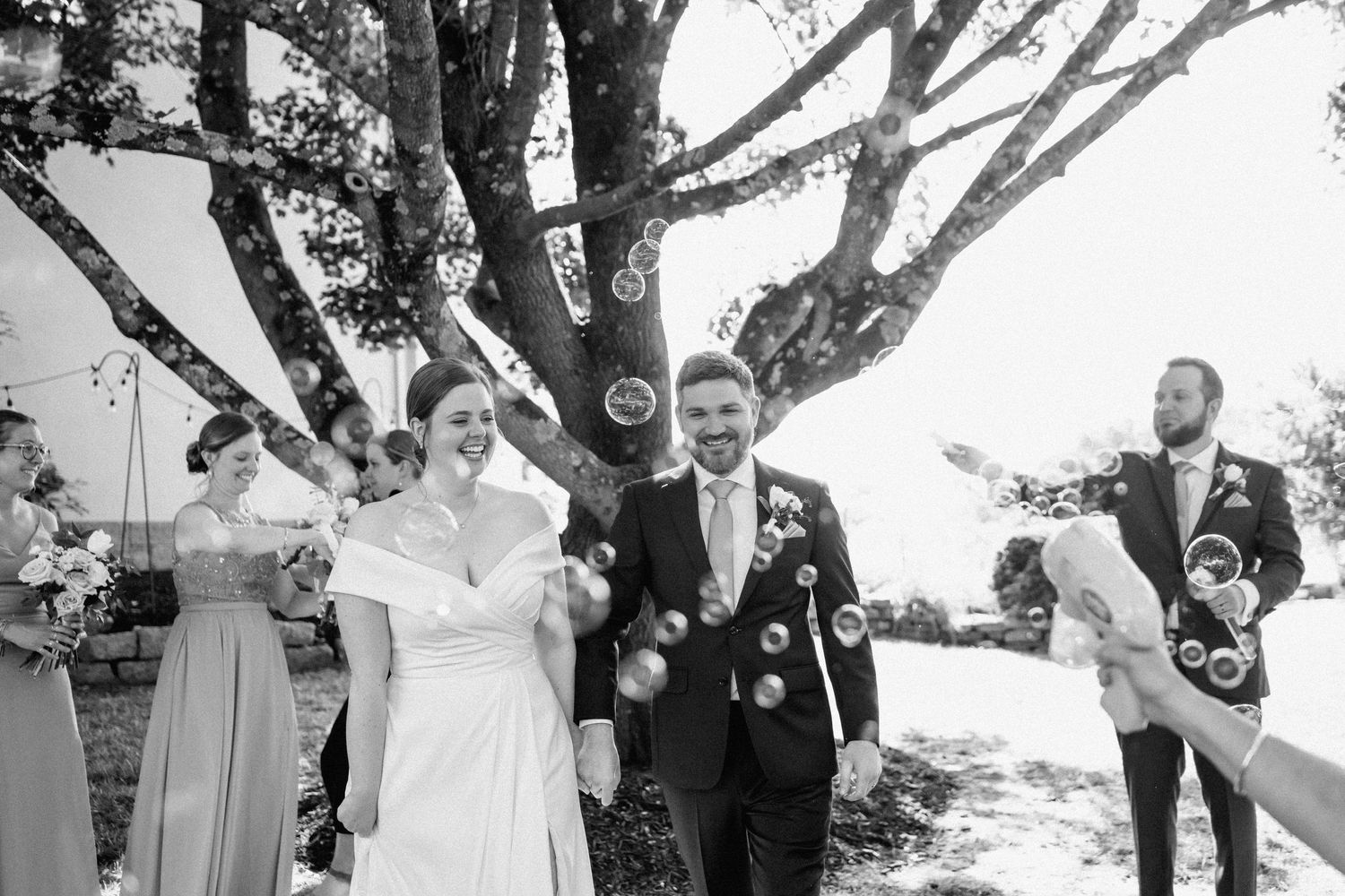 Wedding party celebrates with bubbles under a large tree during outdoor ceremony.