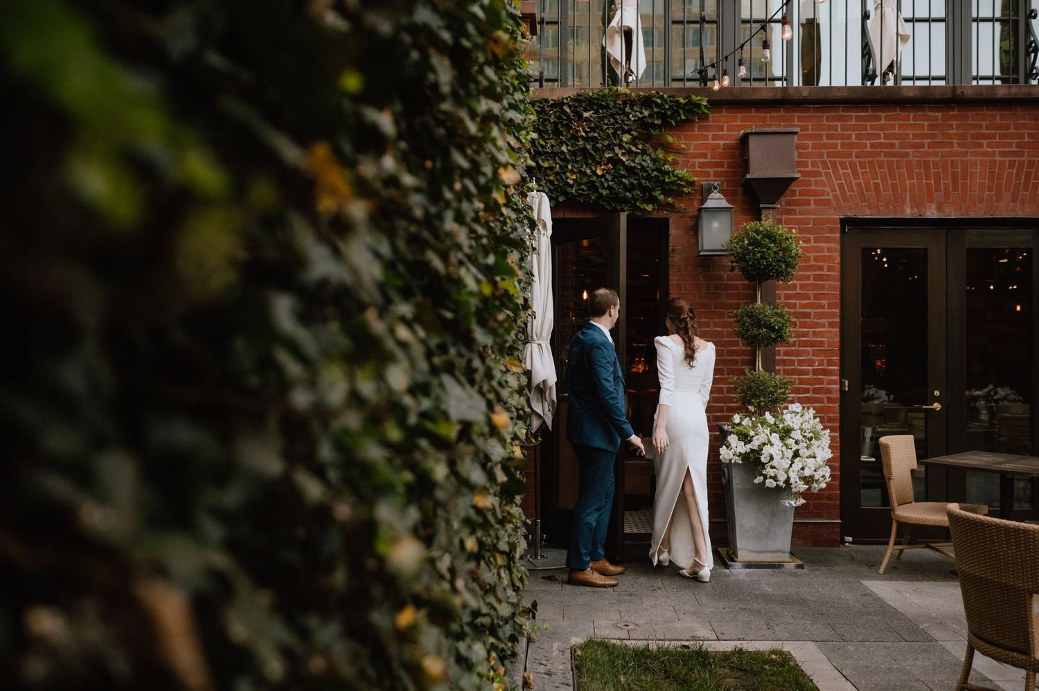 A couple in formal attire stands in a garden courtyard with ivy-covered walls and potted plants.
