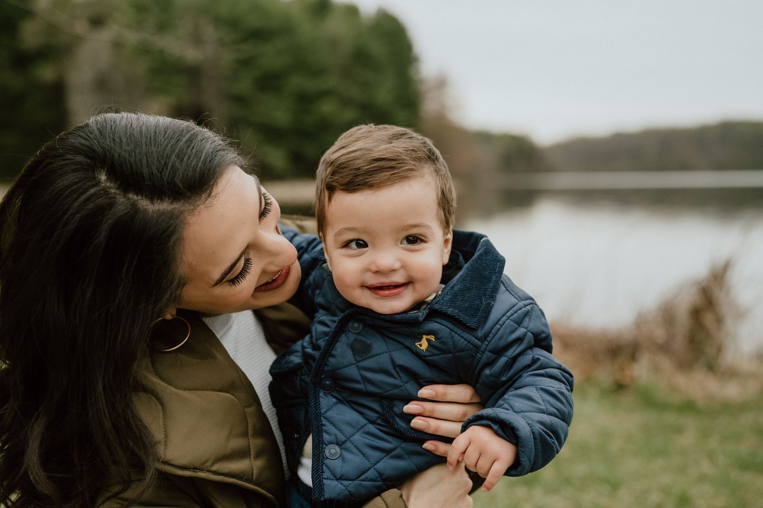 Parent and toddler share intimate moment outdoors with lake in background.