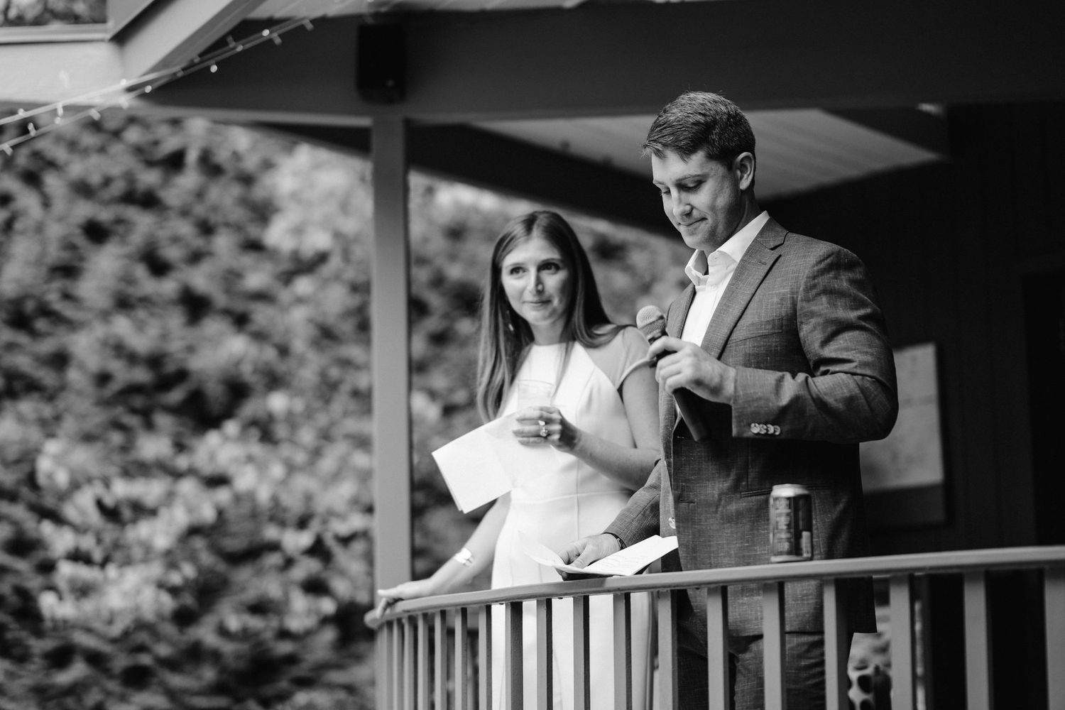 Two people reading from papers while standing on a wooden deck.