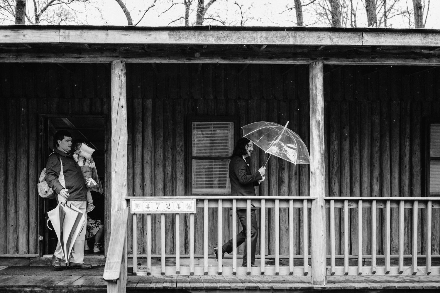 Two figures with umbrellas stand on a rustic wooden porch in black and white.