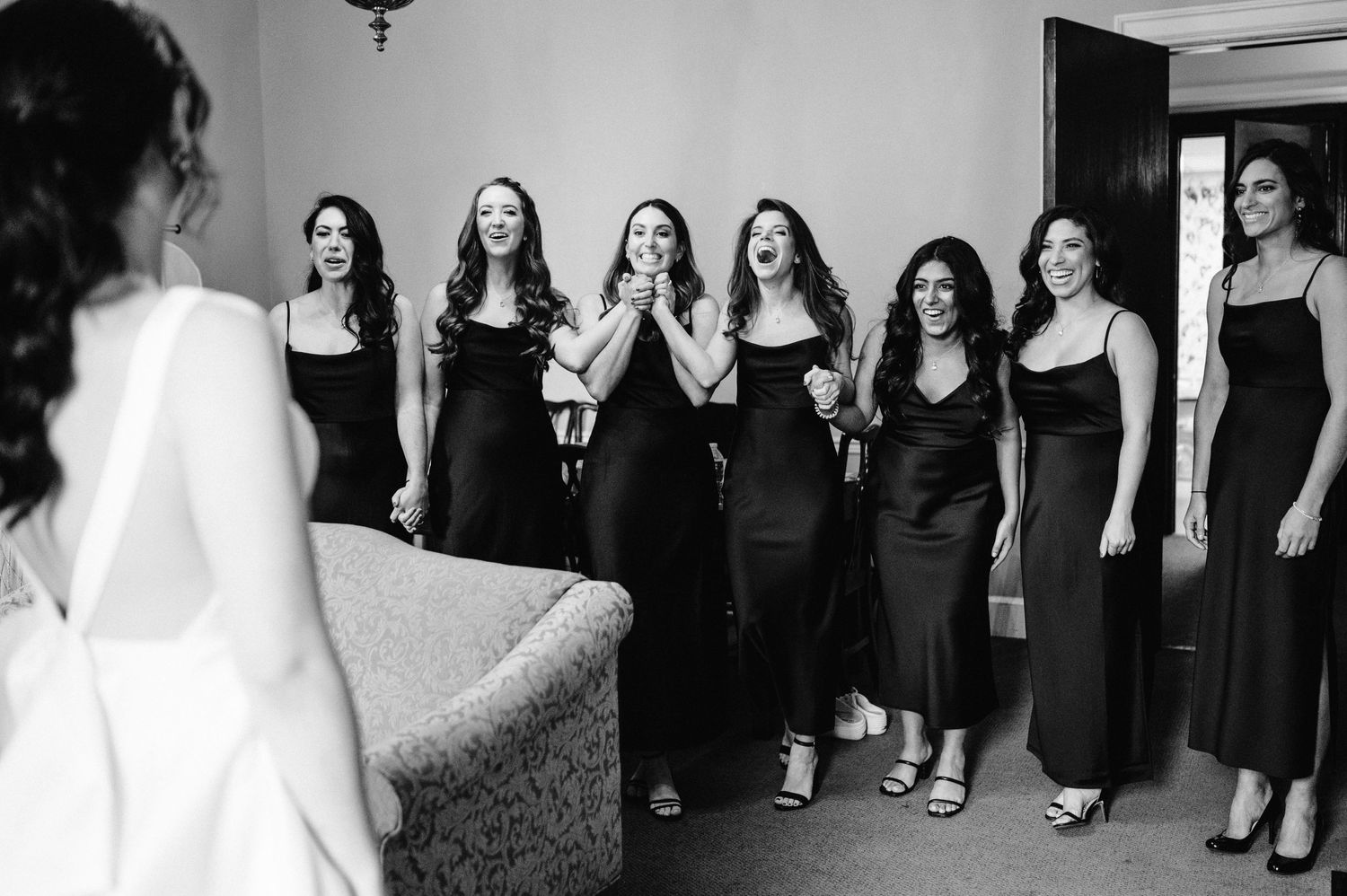Wedding party in matching black dresses reacts with joy to seeing the bride.