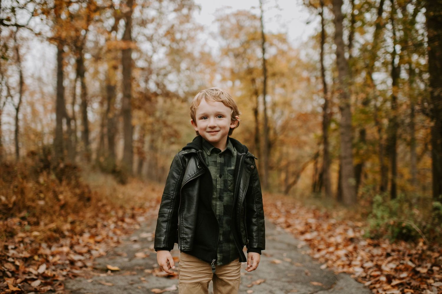 Young child standing alone on forest path wearing black jacket against autumn colors.
