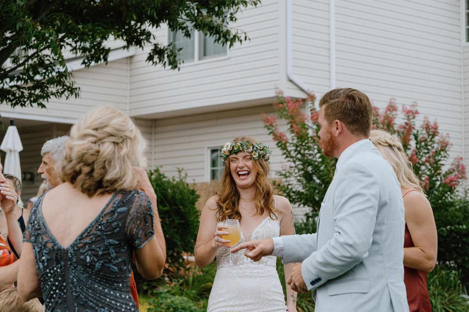 An outdoor wedding celebration with guests gathered under flowering trees and a white house backdrop.