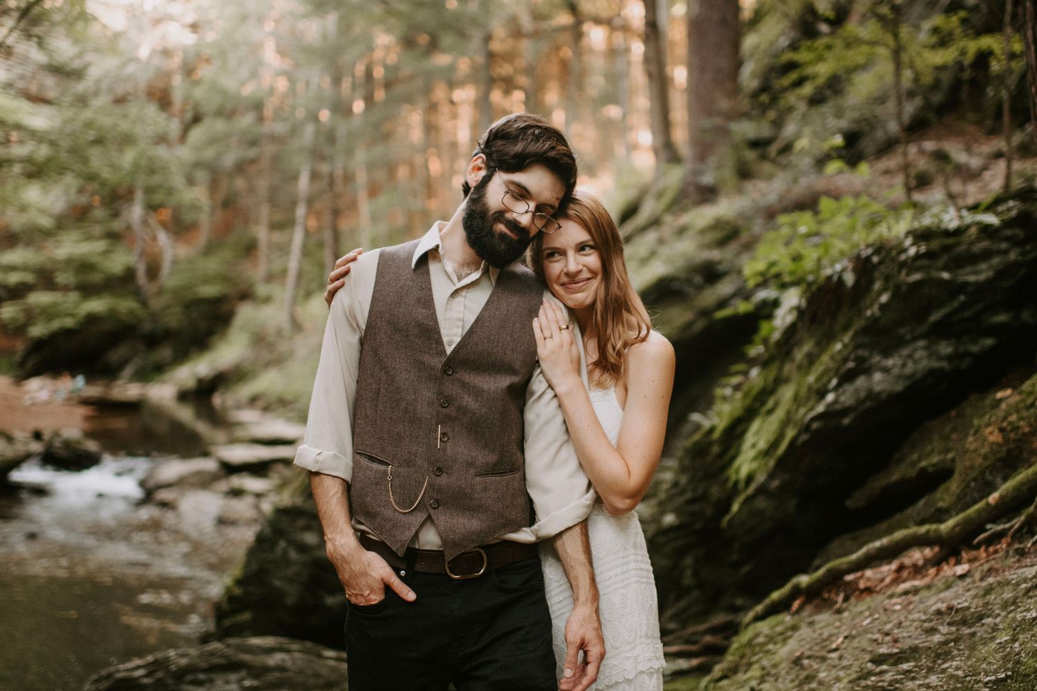 Couple poses together in a scenic forest setting with a stream in the background.