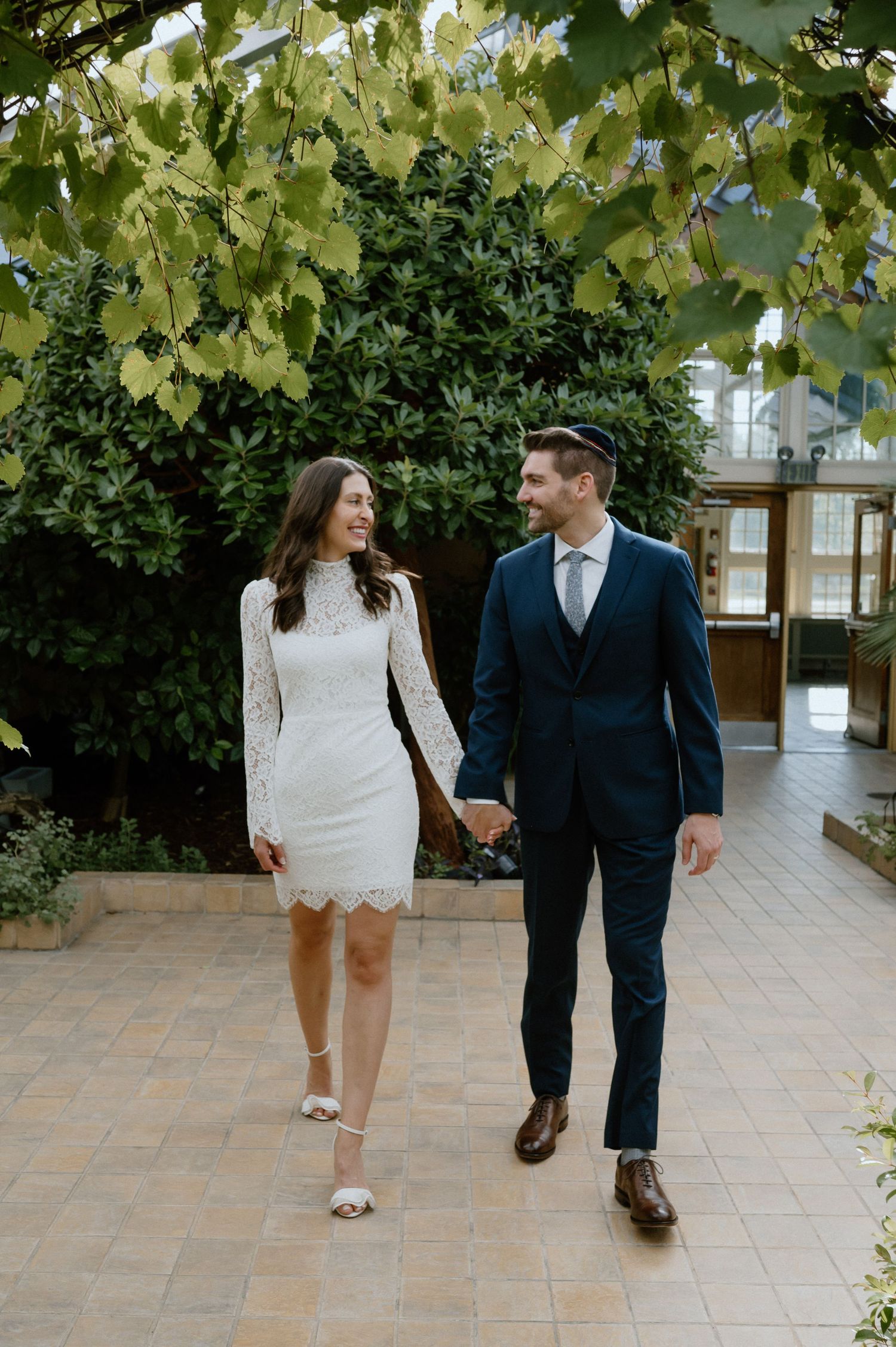 Couple walking hand in hand through garden pathway in wedding attire.