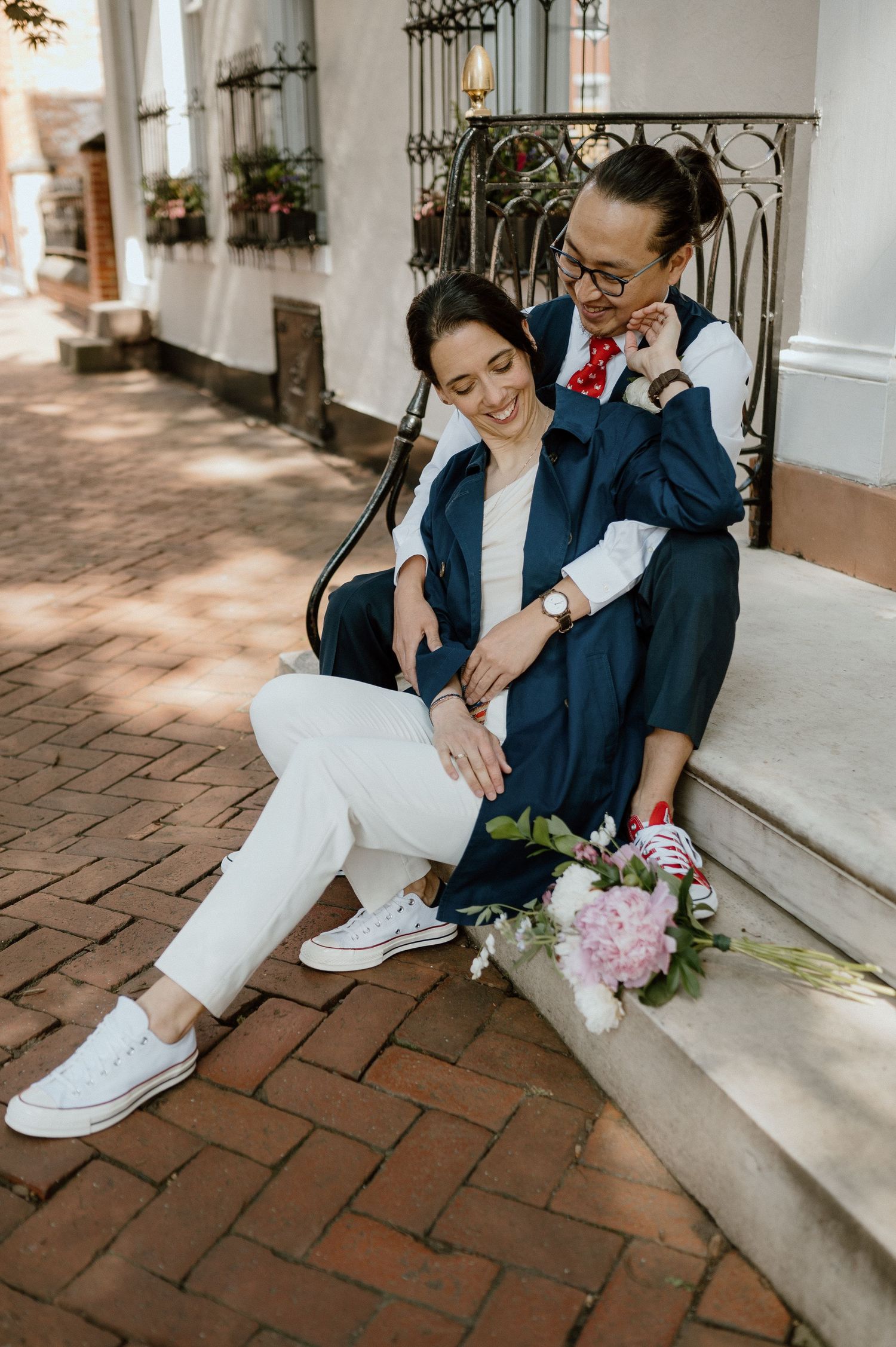 A couple in casual wedding attire sits on stone steps with flowers in a romantic pose.