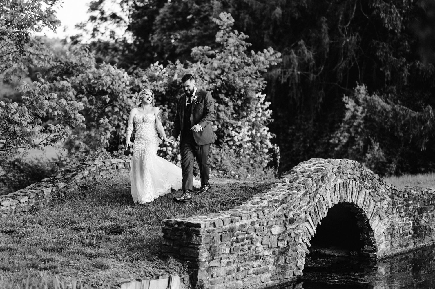 Black and white photo of newlyweds standing on historic stone bridge in garden setting.