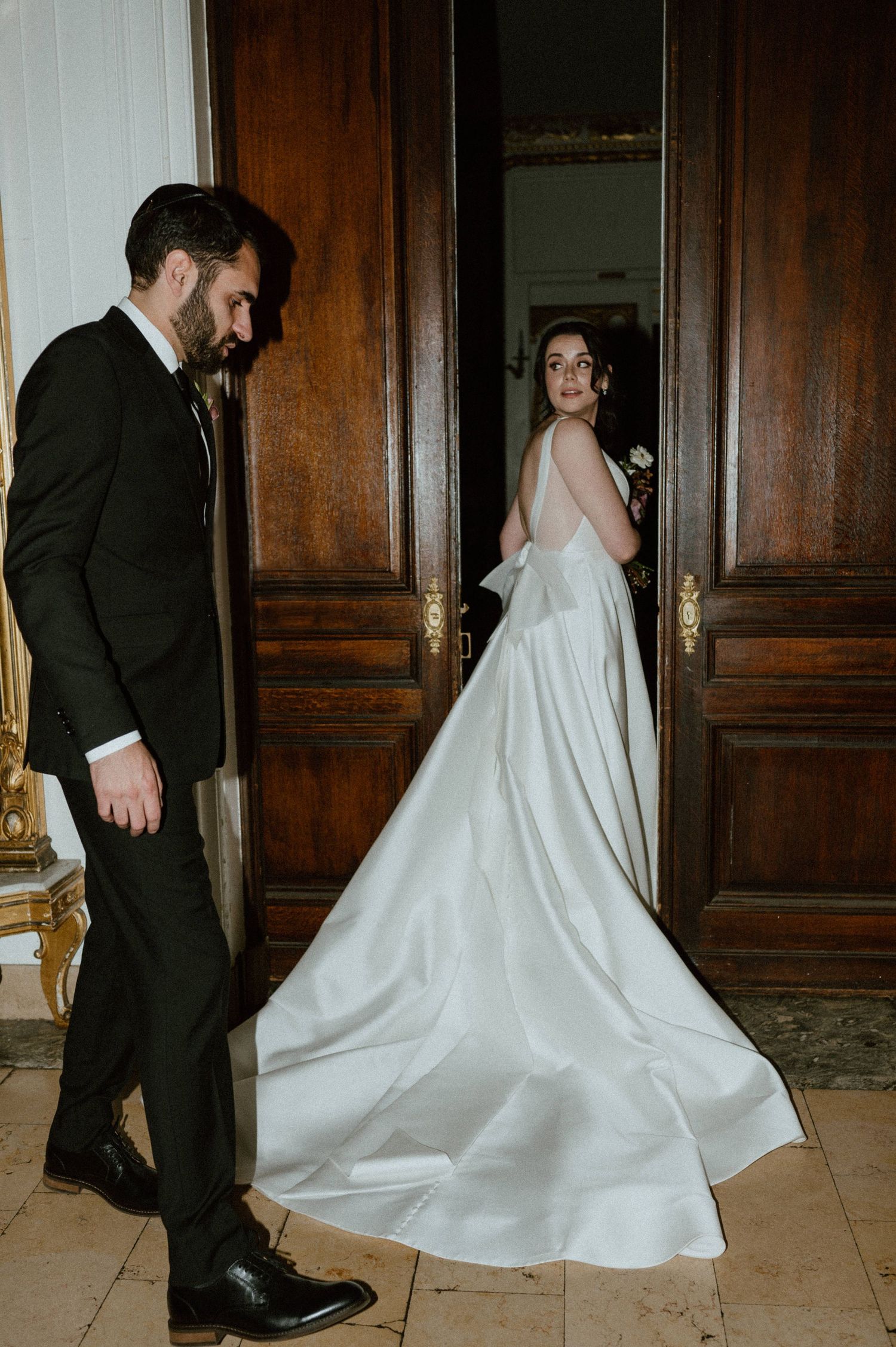 Elegant bride in flowing white dress looking over her shoulder in a grand hallway with ornate wooden doors.