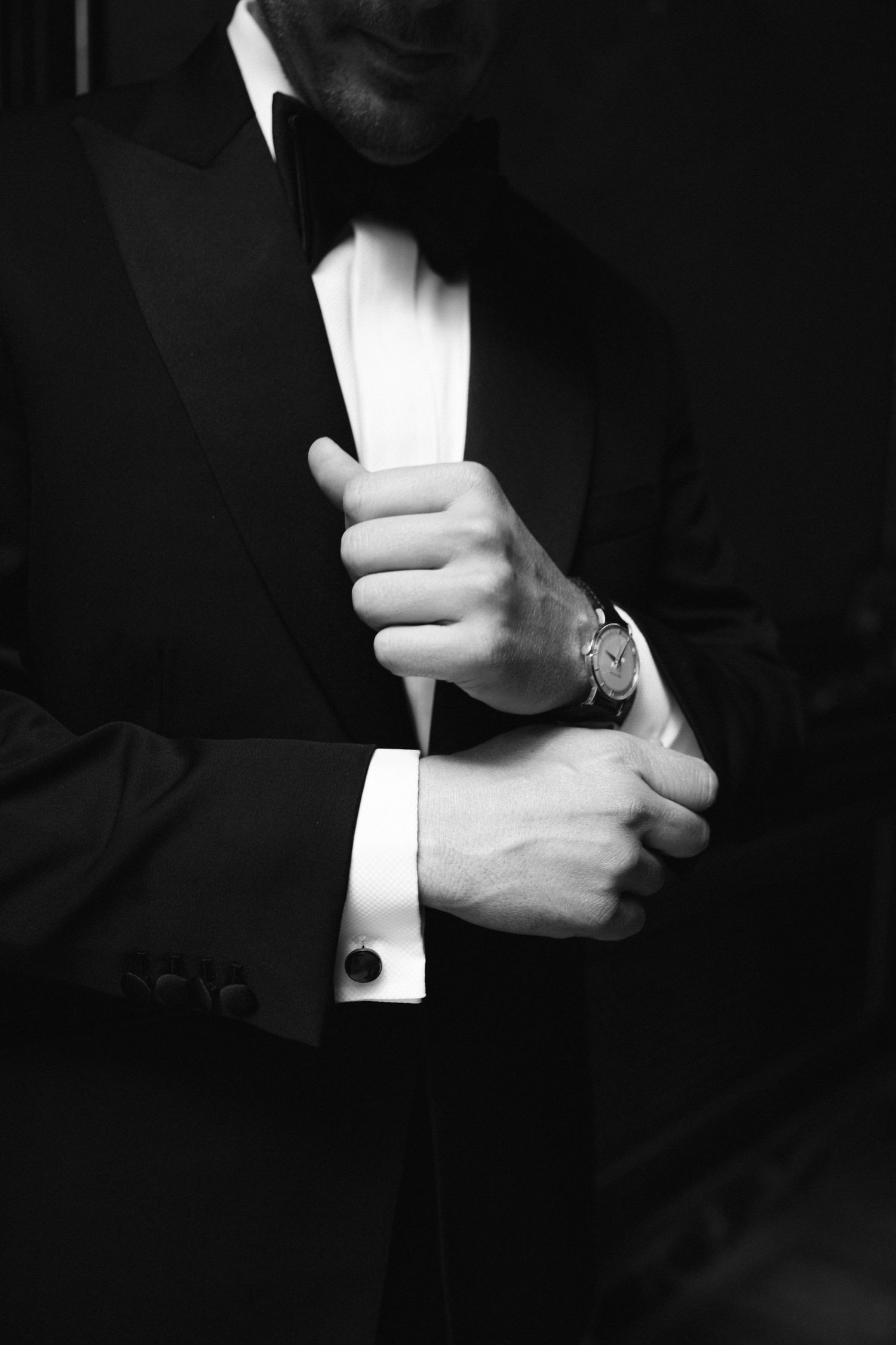 Close-up of grooms hands adjusting tuxedo cuffs and sleeve showing elegant watch detail in black and white.