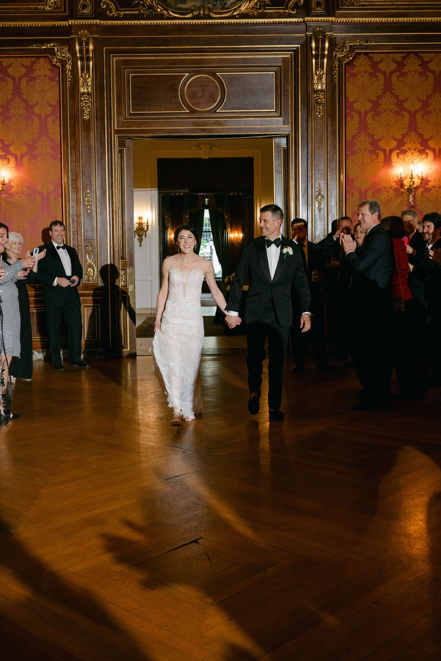 A couple walks across a grand ballroom with ornate gold trim and wall sconces during an elegant evening celebration.