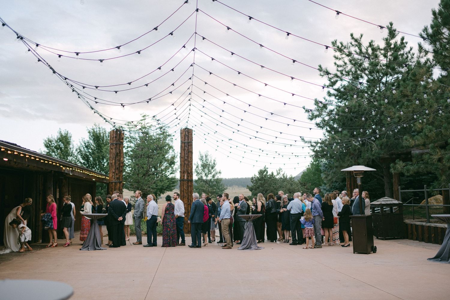 Wedding guests gather under string lights at an outdoor reception venue.