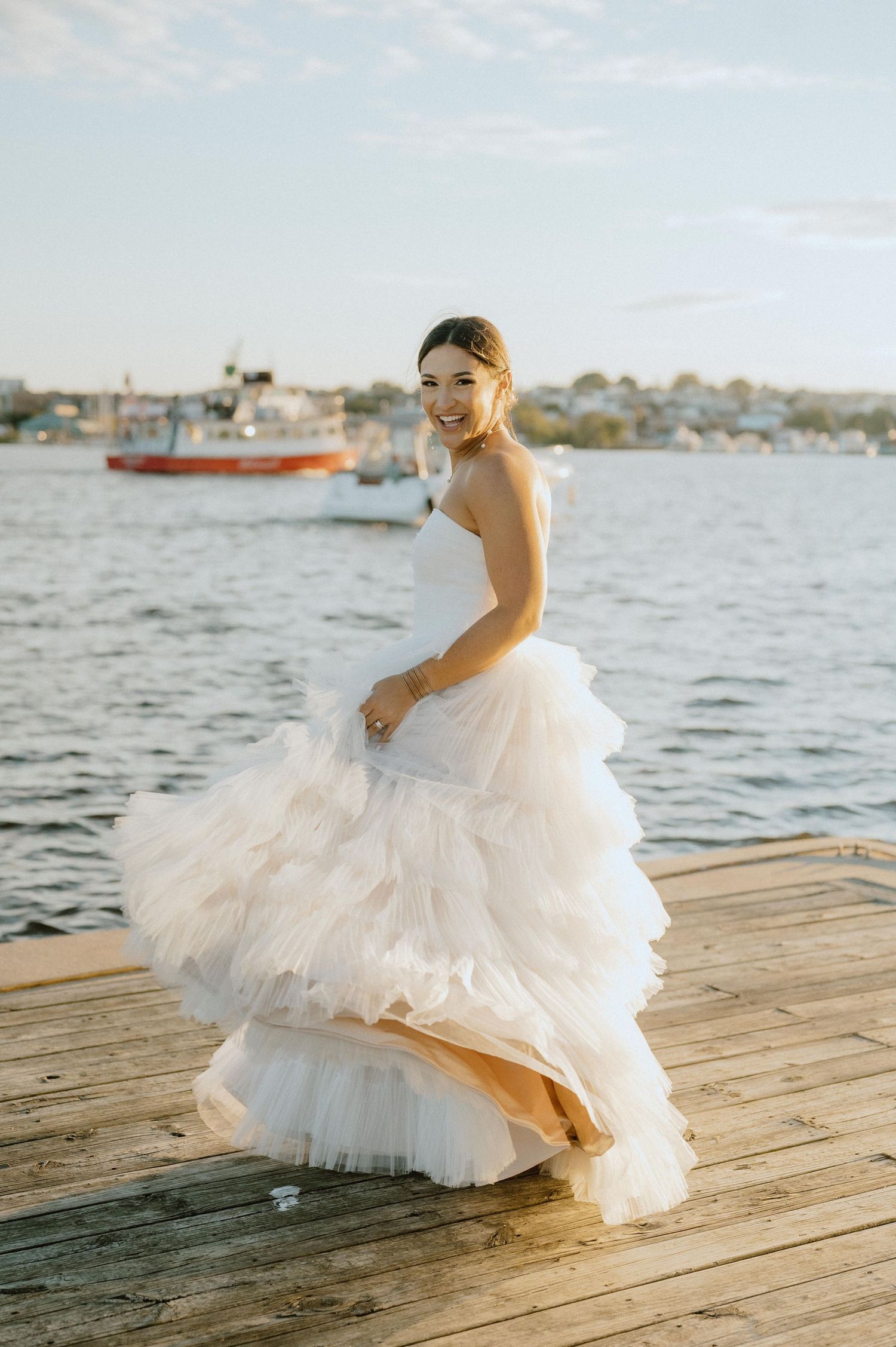 Bride in layered tulle wedding gown poses on wooden dock with harbor in background.