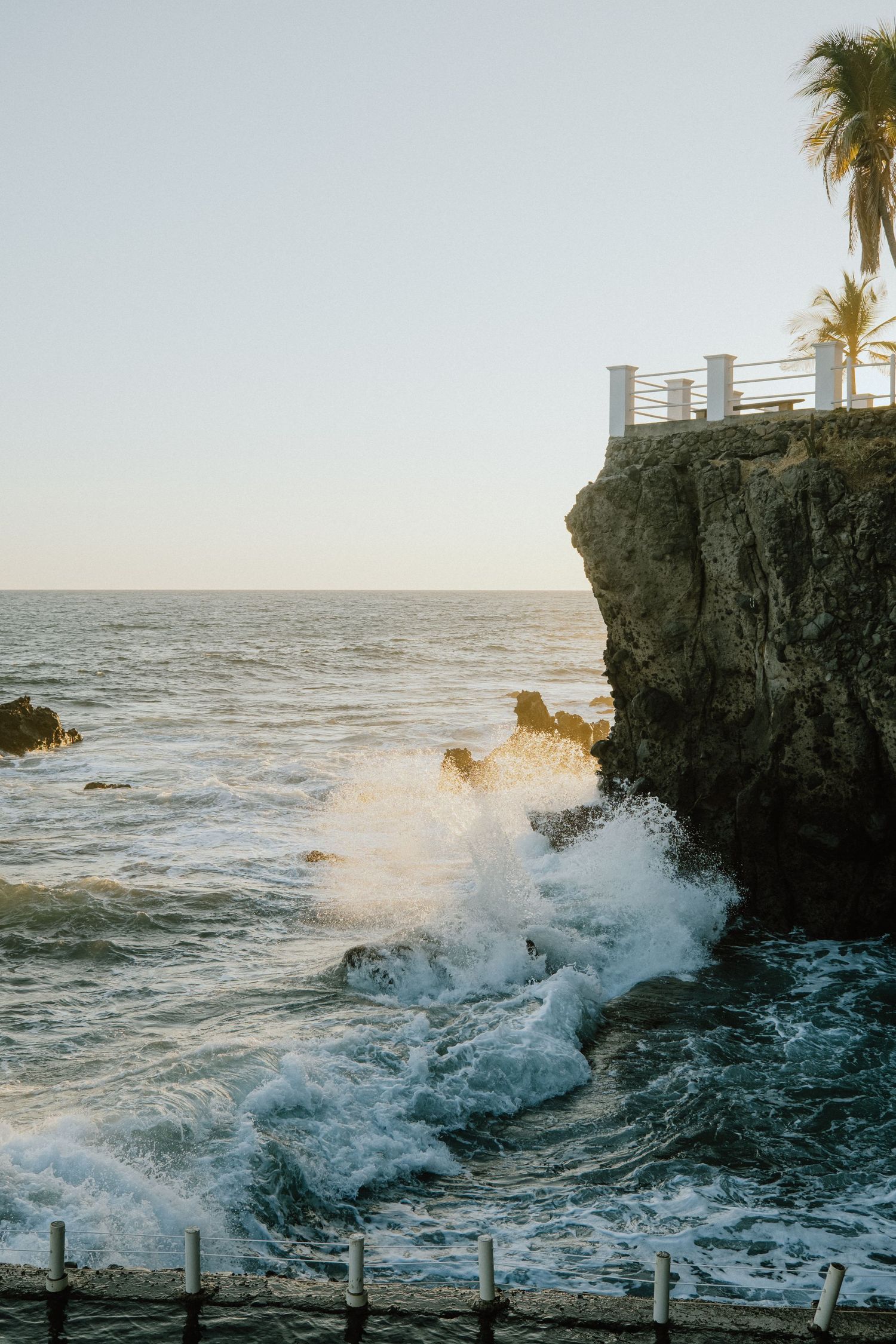 Dramatic waves crash against coastal cliffs with palm trees and white railings visible at sunset.
