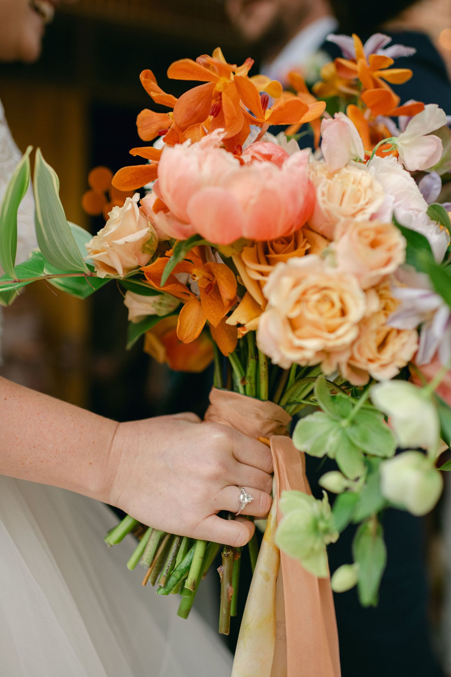 A stunning wedding bouquet with orange lilies, peach roses, and greenery tied with silk ribbon.