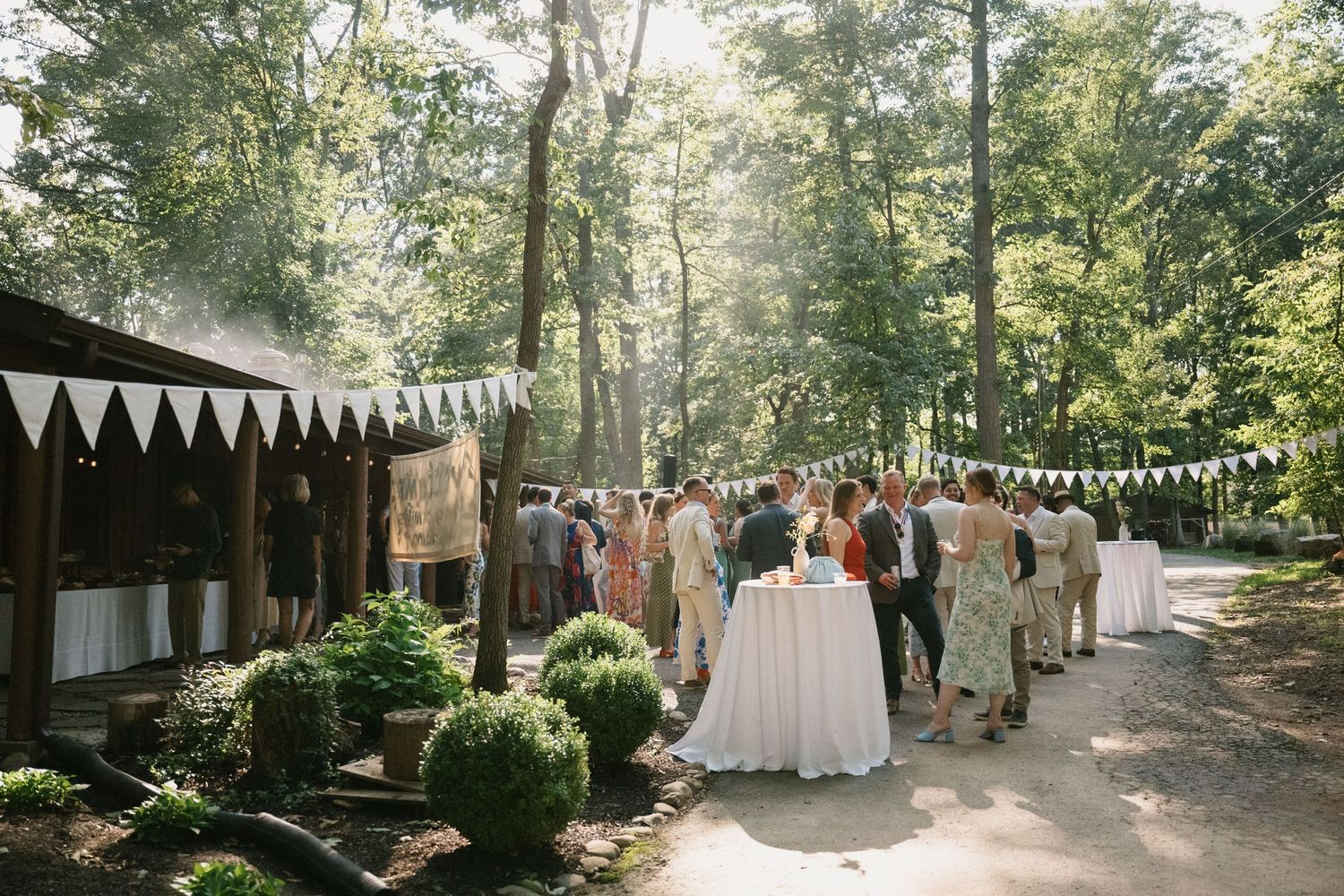 Guests mingle at an outdoor wedding reception with white bunting decorations and tall trees.