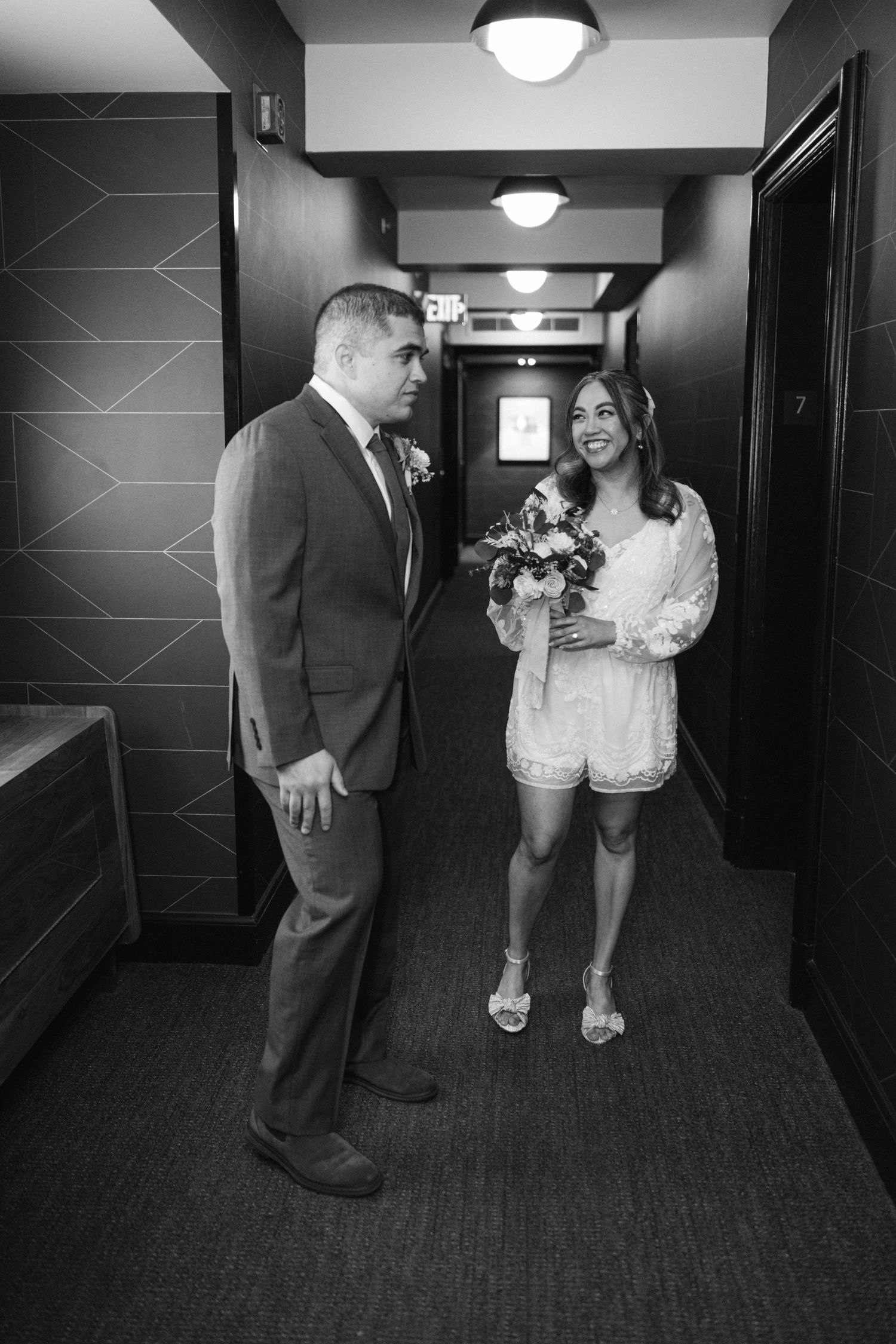 A couple shares a joyful moment in a hotel hallway wearing formal attire and holding a bouquet.