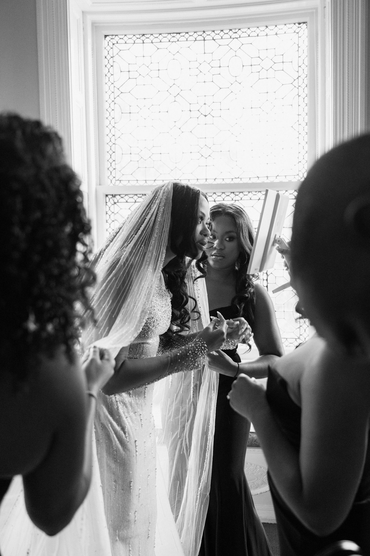 Silhouetted figures help adjust a wedding veil near a bright window.