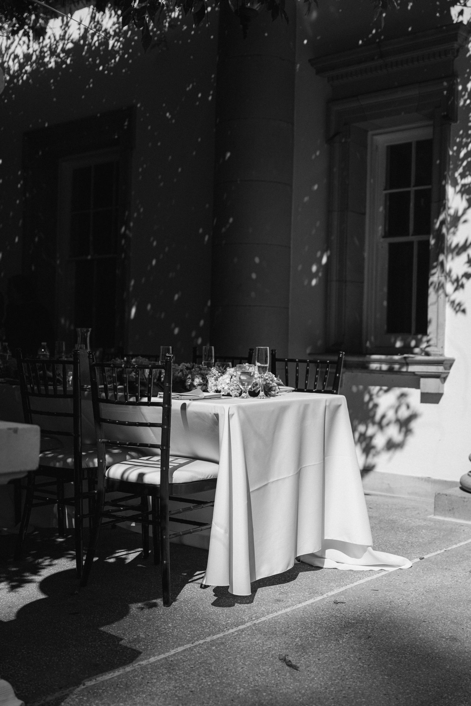 Black and white photo of outdoor reception area with round tables and chairs casting dramatic shadows.