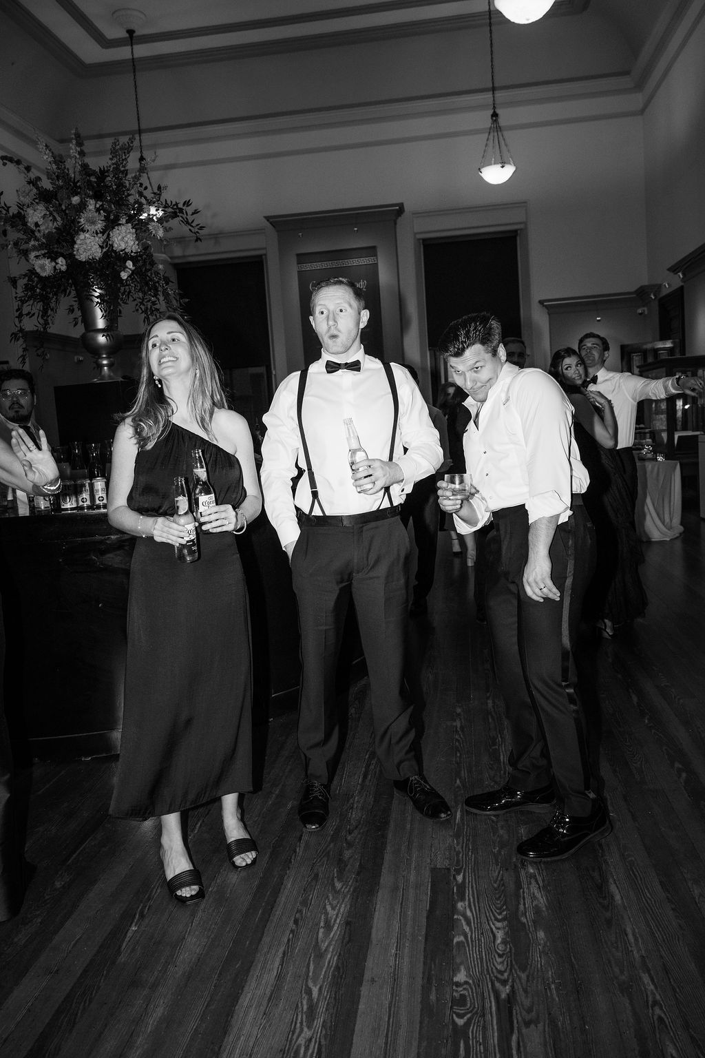 Wedding guests in formal attire dancing on wooden floor under pendant lights.