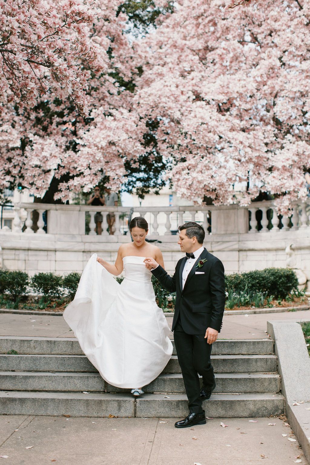 Bride in white gown descends stone steps with groom under cherry blossoms.