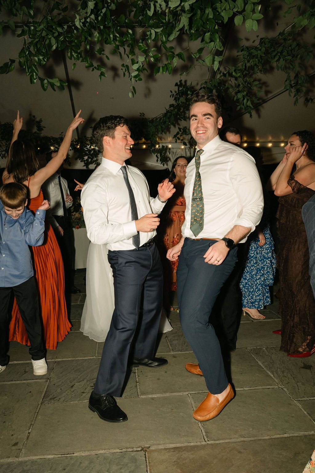 Two guests dancing and enjoying themselves at a wedding reception under string lights and greenery.