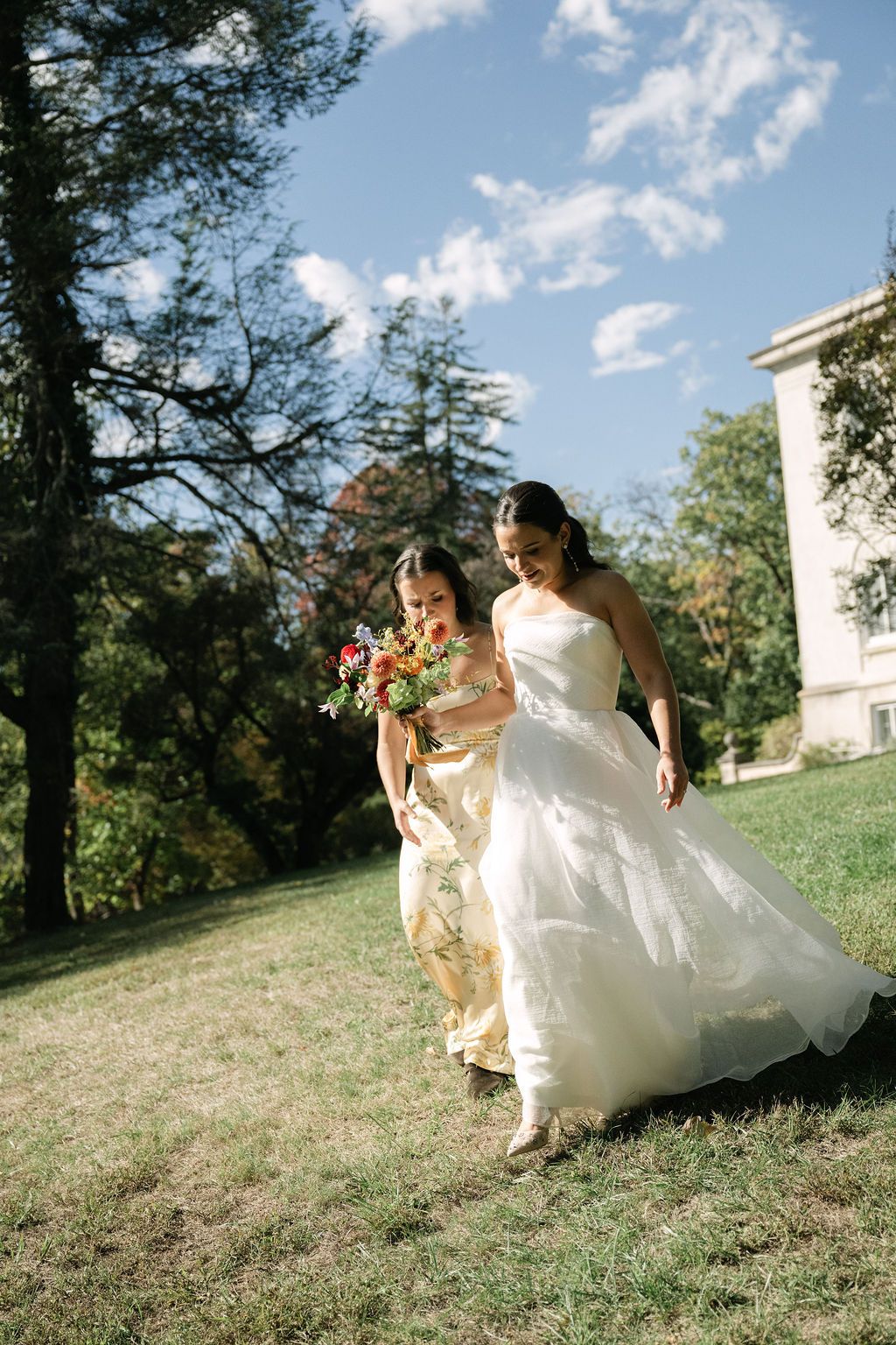 People in formal wedding attire run playfully down a grassy hill on a sunny day.