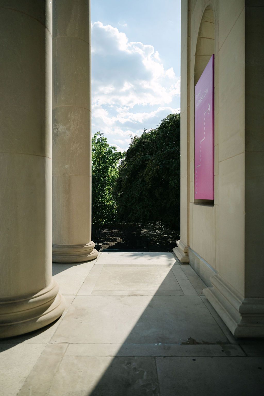 Architectural columns frame a view of trees and sky through a classical colonnade walkway with pink panels.