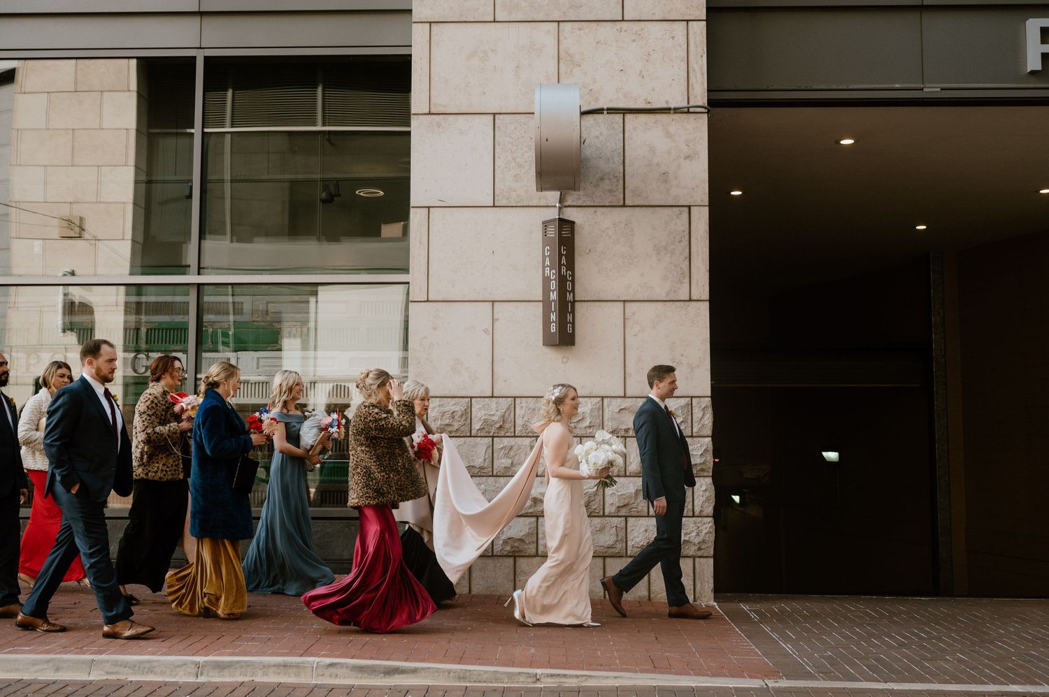 Wedding party walking along stone building exterior in formal attire with flowing dresses.